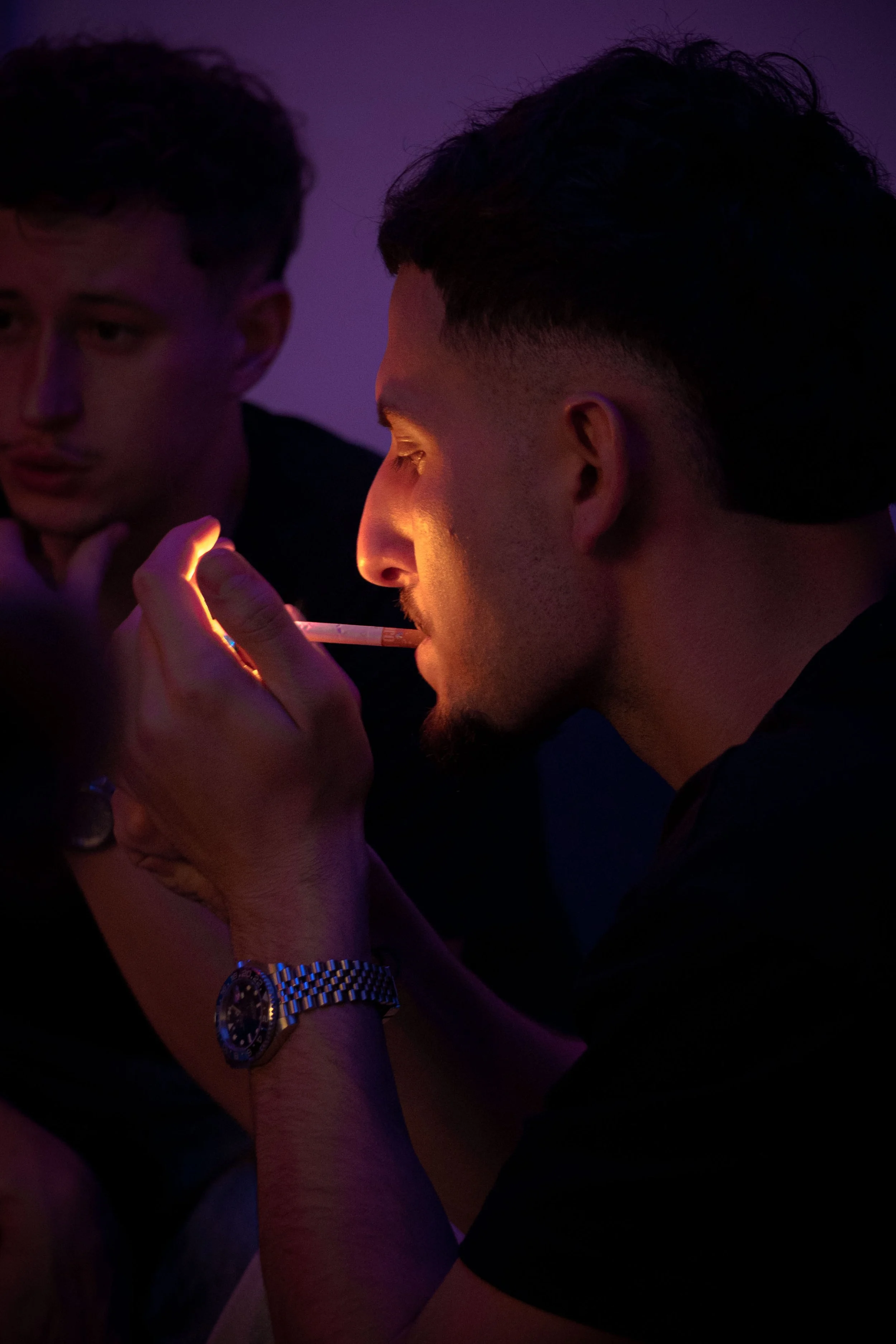 Deux jeunes hommes sont proches, l'un d'eux fume une cigarette, dans une lumière tamisée violette et orange.