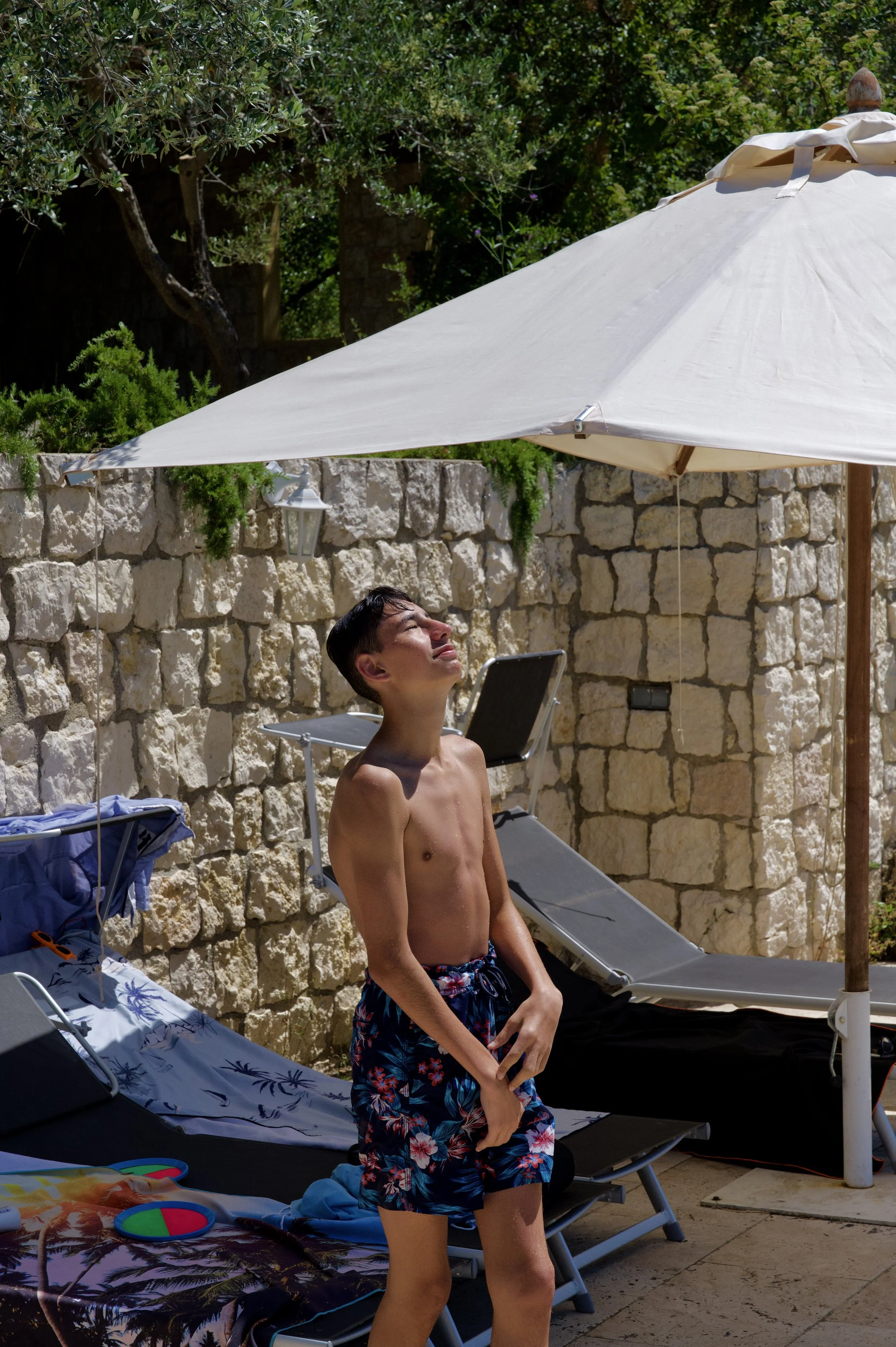 Jeune garçon en maillot de bain à fleurs, debout sous un parasol blanc, au bord d'une piscine, avec un mur en pierre et des arbres en arrière-plan.