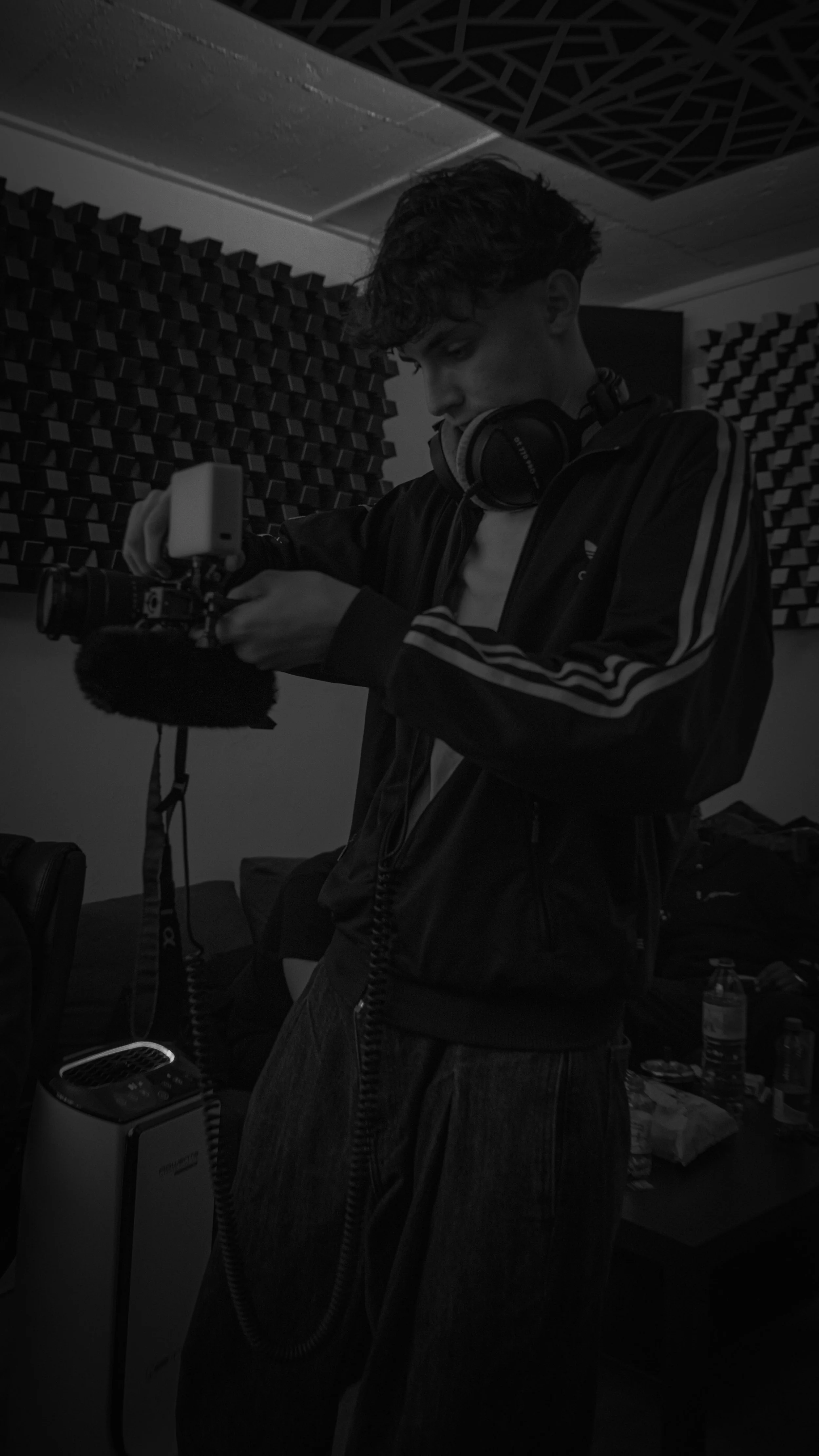 Un jeune homme avec des écouteurs portant une veste à rayures, tenant un appareil dans un studio d'enregistrement, avec des panneaux acoustiques sur le mur.