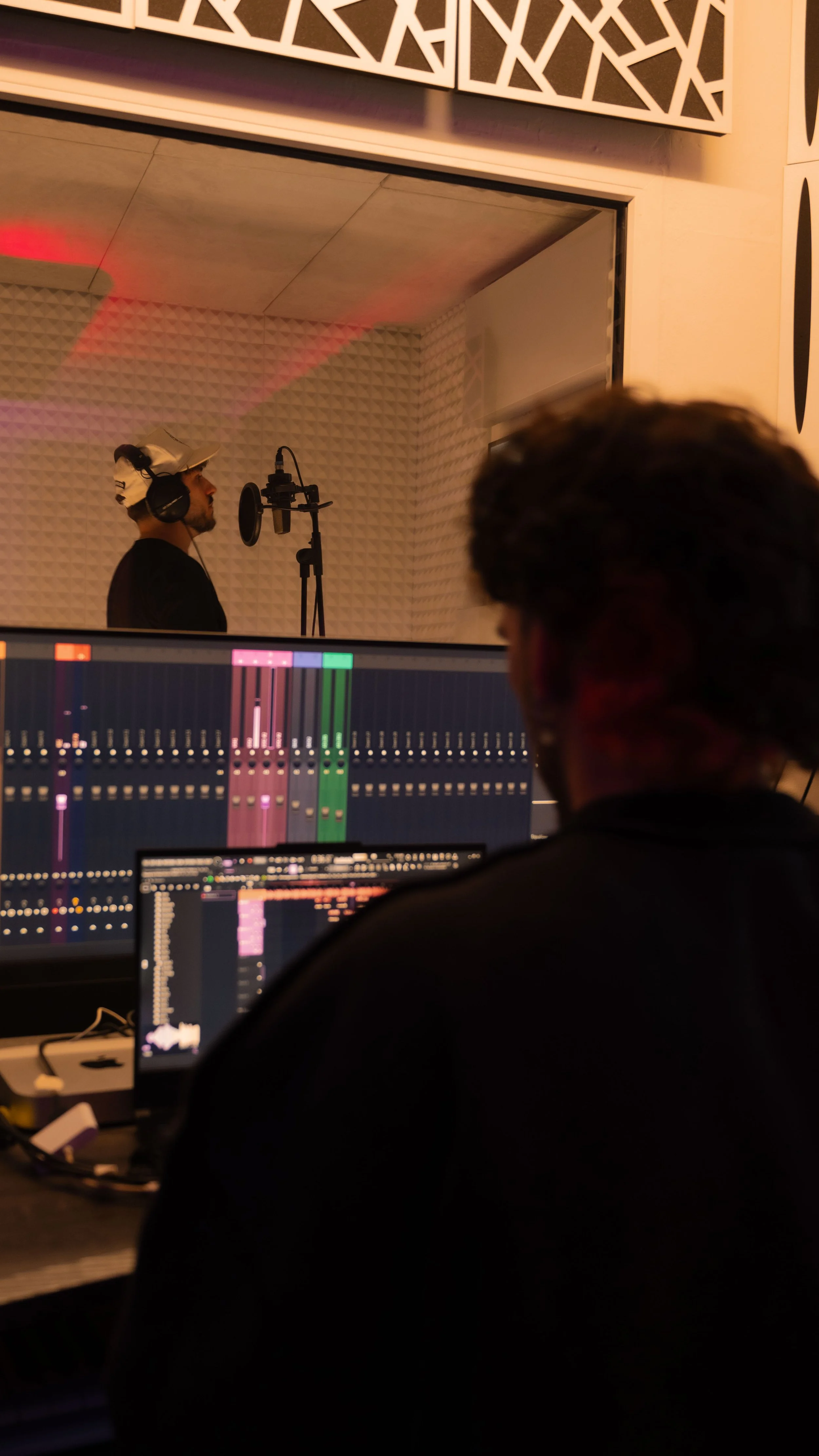 Une personne dans une cabine d'enregistrement chantant ou enregistrement vocal, un microphone, un casque audio, et un ingénieur du son ou producteur travaillant sur un ordinateur avec un logiciel de production musicale dans une pièce insonorisée.