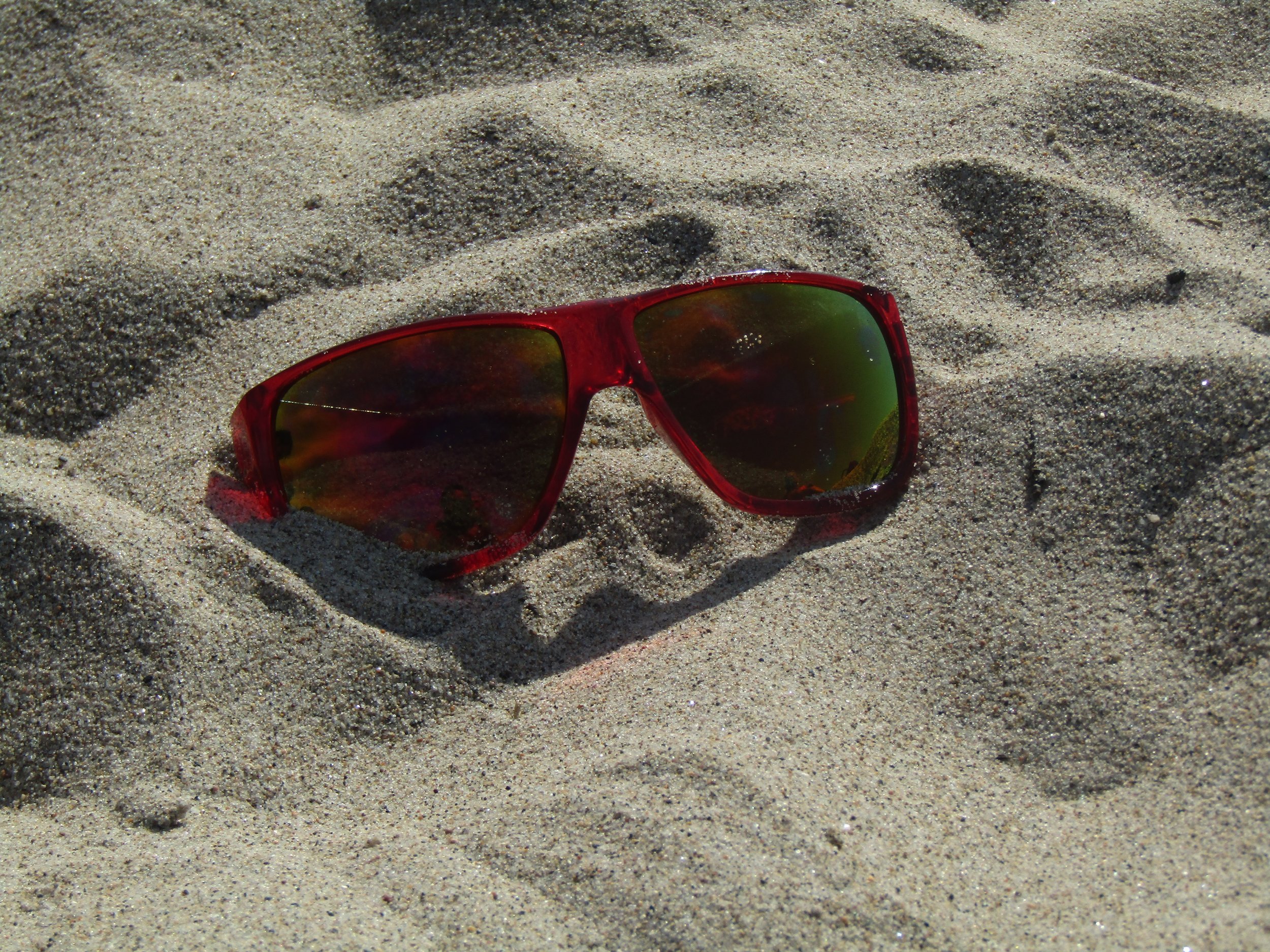 Red sunglasses with reflective green lenses resting on sandy beach.