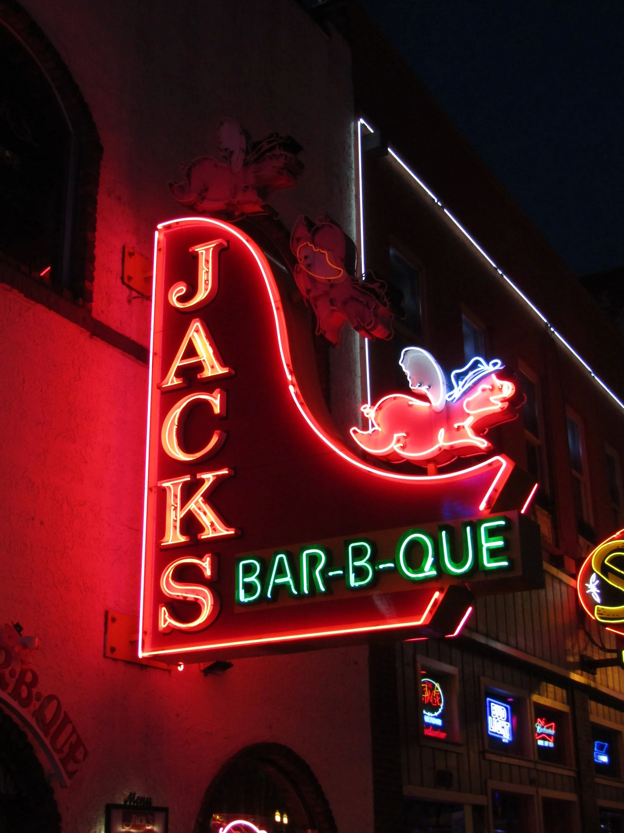 Neon sign for Jack's Bar-B-Que featuring a red and pink mermaid riding a dragon, with the text 'JACK'S' in vertical red neon and 'BAR-B-QUE' in green neon below.