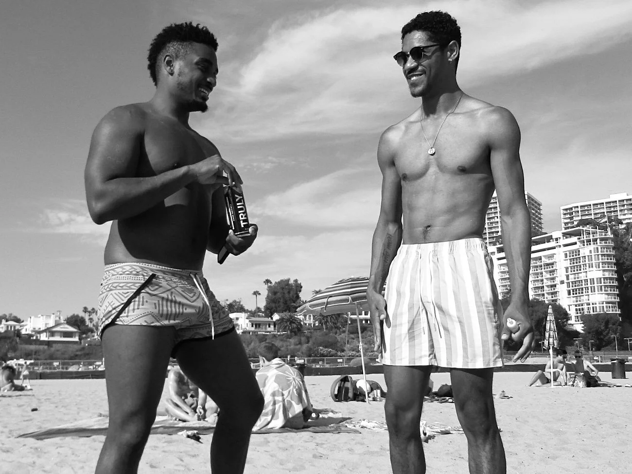 Two shirtless men standing on a beach, smiling and engaging in conversation, with umbrellas, beachgoers, and high-rise buildings in the background.