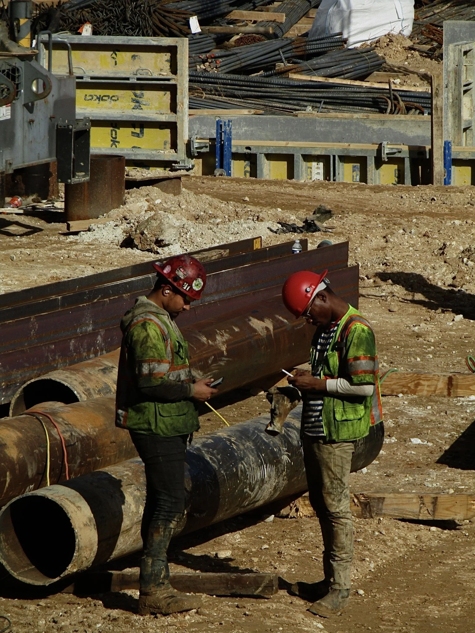 Two construction workers with red safety helmets and green reflective vests standing on dirt at a construction site with large pipes and metal framework in the background, both looking at their phones.