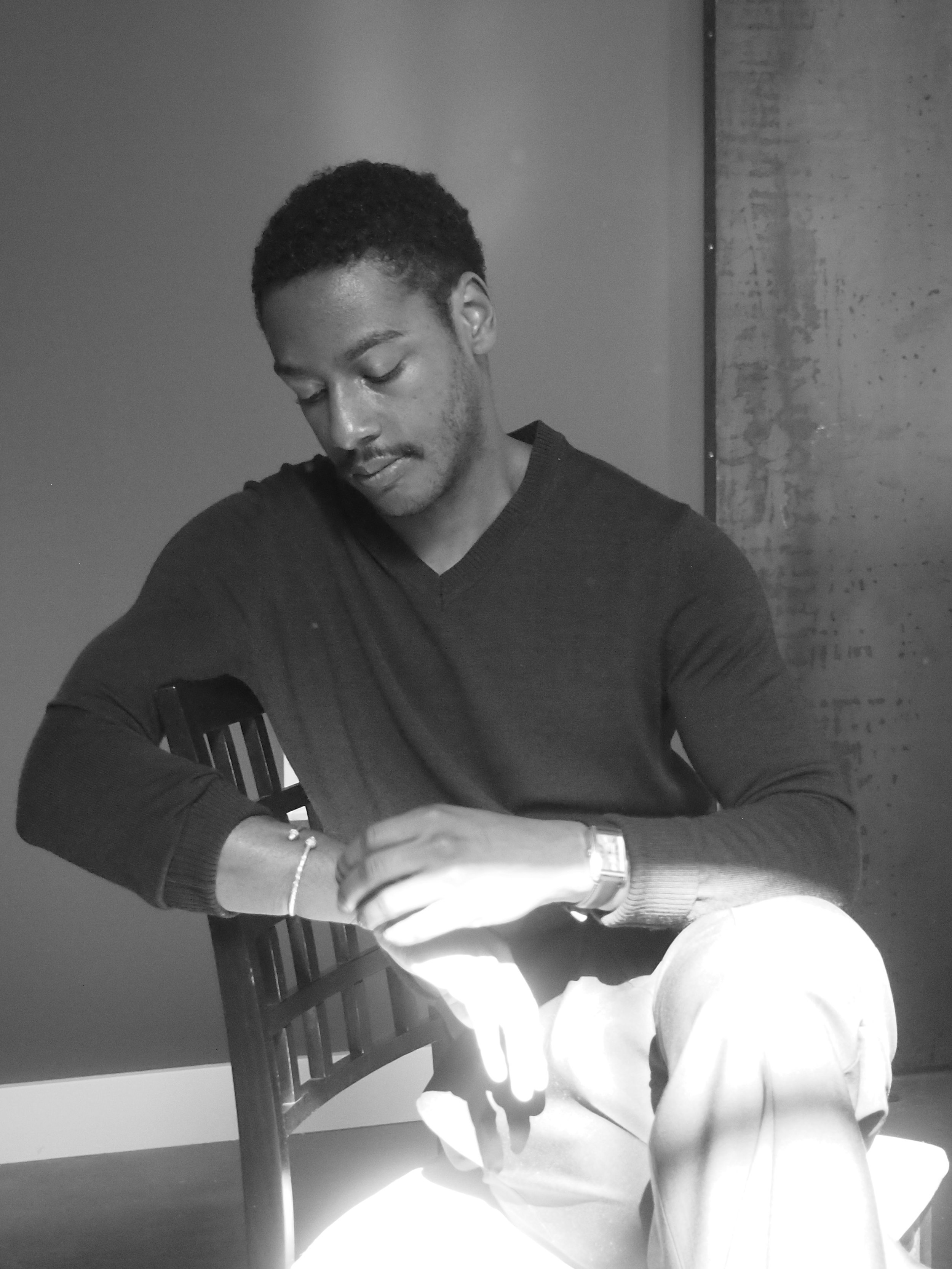 A young man sitting on a wooden chair, looking down at his left wrist. He has short curly hair, a slight mustache, and is wearing a long-sleeve dark sweater, light-colored pants, a wristwatch on his left wrist, and a bracelet on his right wrist. The room has a concrete wall and a smooth wall divided by a vertical line.