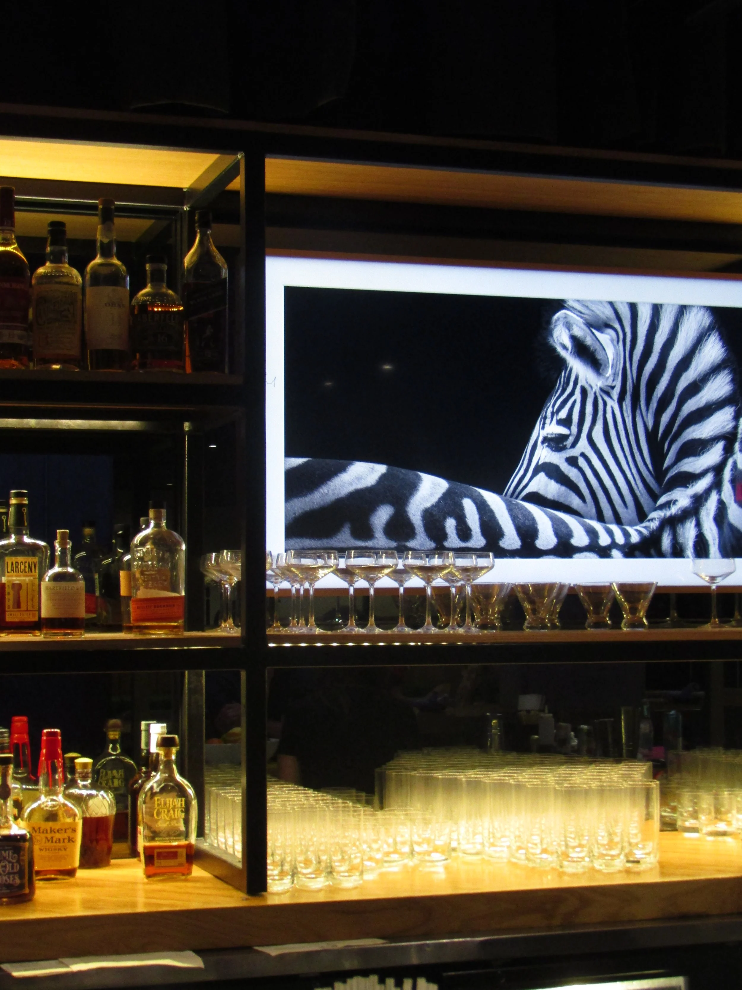 Bar shelf with various liquor bottles, a row of empty cocktail glasses, and a large screen displaying a black-and-white image of a zebra.