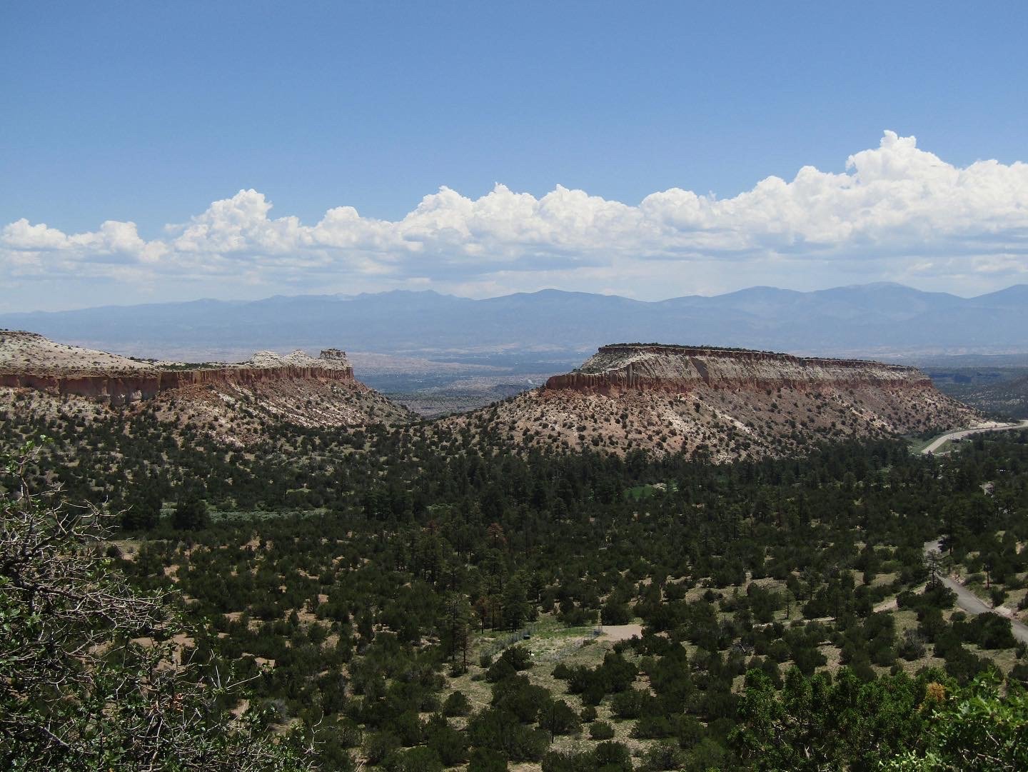 Scenic landscape with layered mesas and hills, sparse green vegetation, a mountain range in the distance, and a partly cloudy sky.