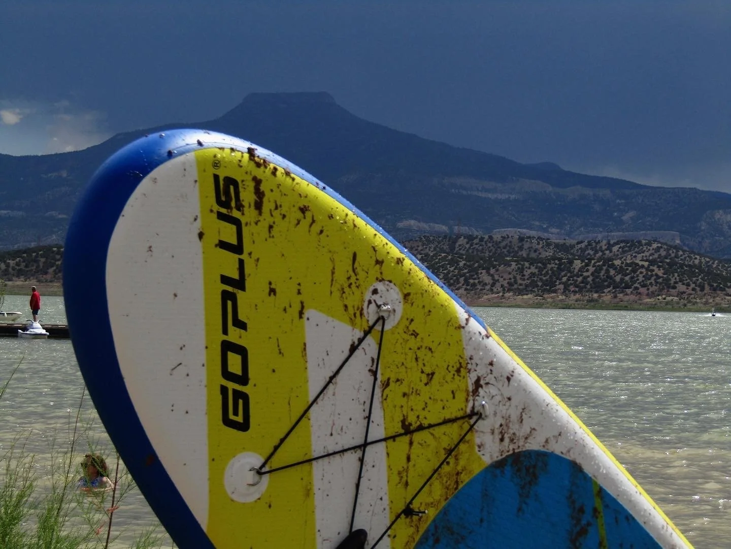 Close-up of a paddleboard with the brand name 'GOPLUS,' wet and slightly dirty, resting on the grass near the water, with a mountain and cloudy sky in the background, and a person standing on a boat in the lake.