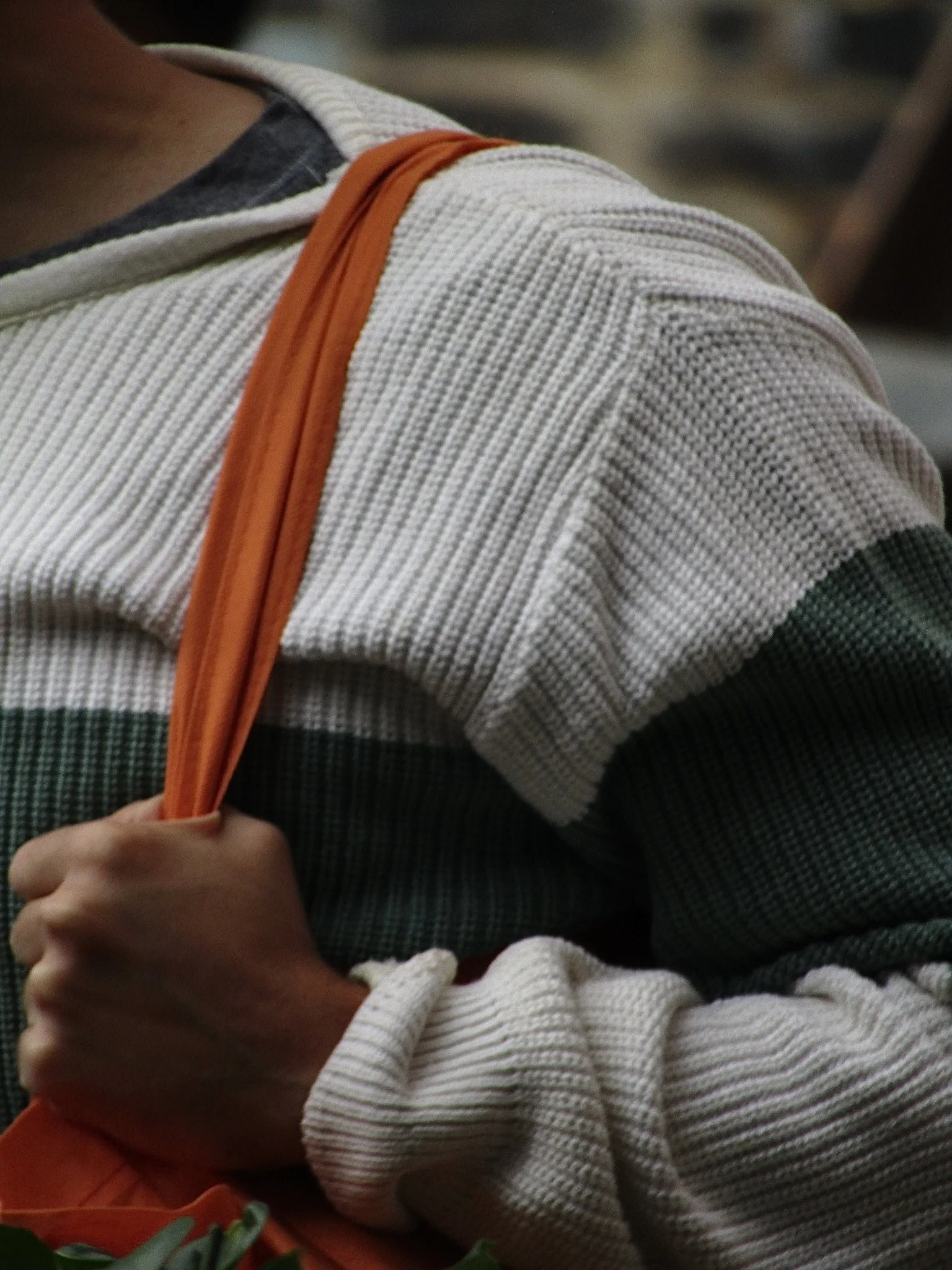 Close-up of a person wearing a multicolored striped sweater with an orange bag strap over the shoulder, holding the strap with their hand.