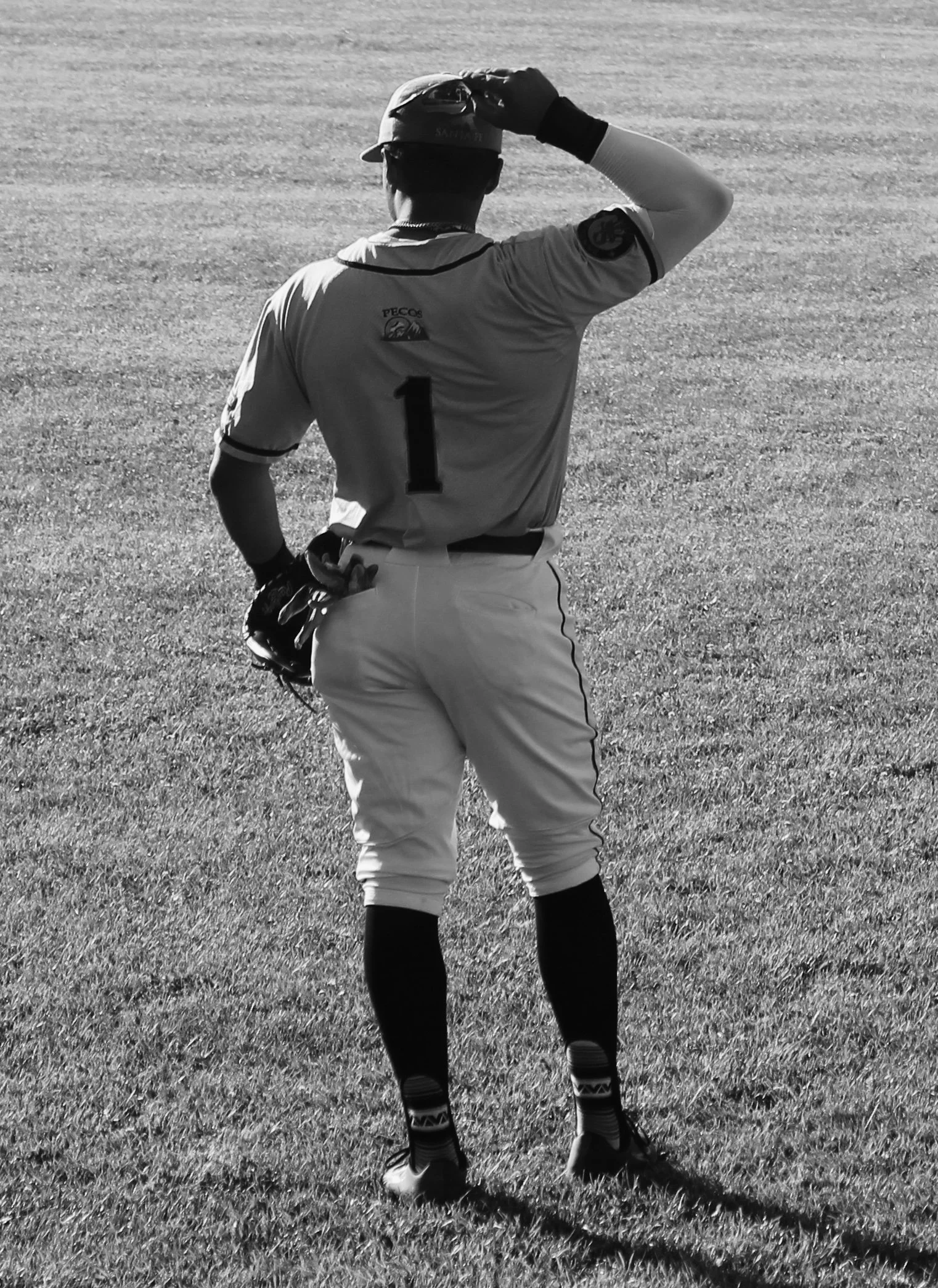 A baseball player in uniform stands on the field, holding a baseball glove in their left hand, with their right hand touching their baseball cap, viewed from the back.