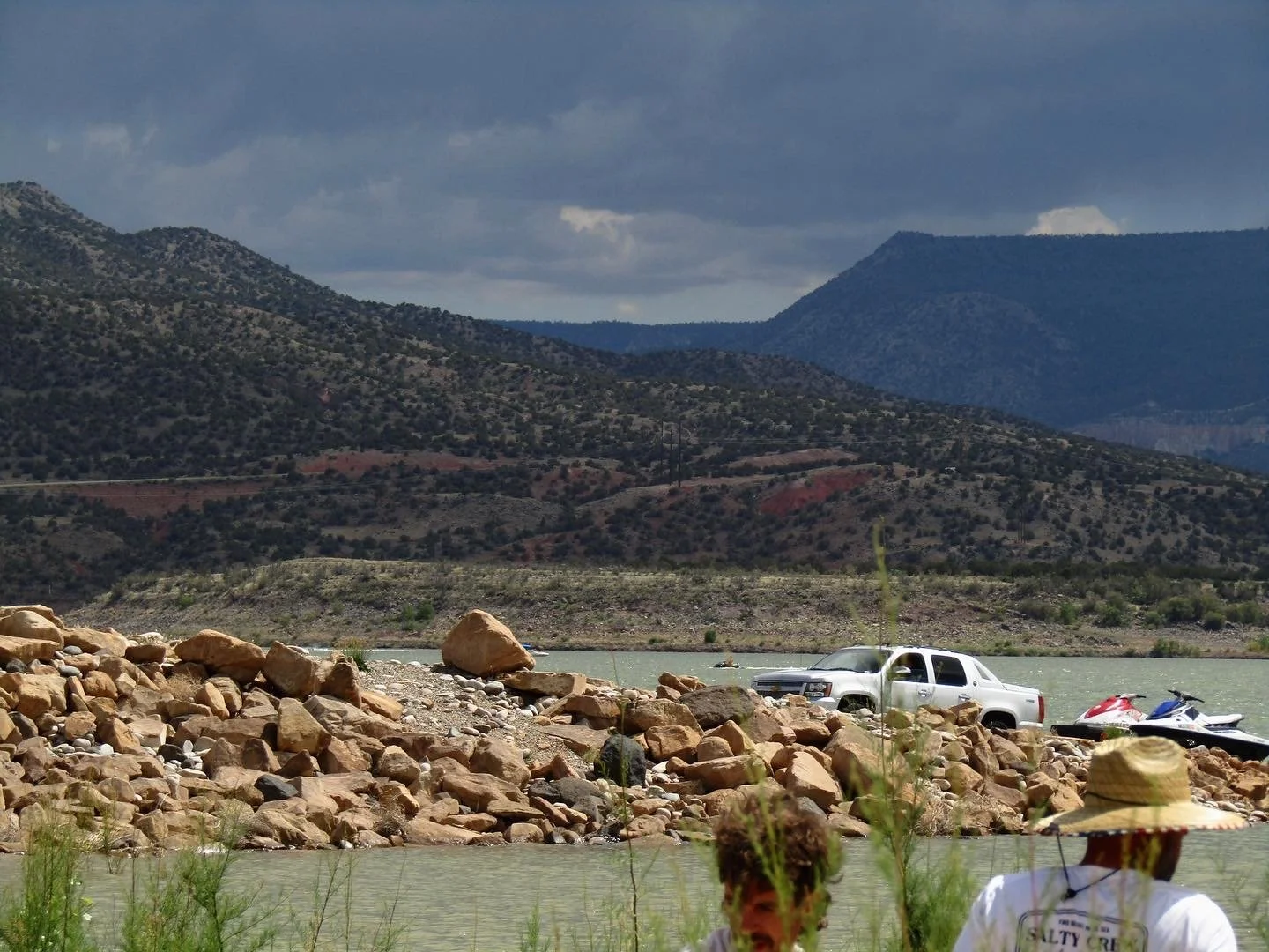 A river or lake with rocky shoreline in a mountainous landscape. Several people are in the water, one wearing a straw hat, and there is a white truck and jet skis on the shore.