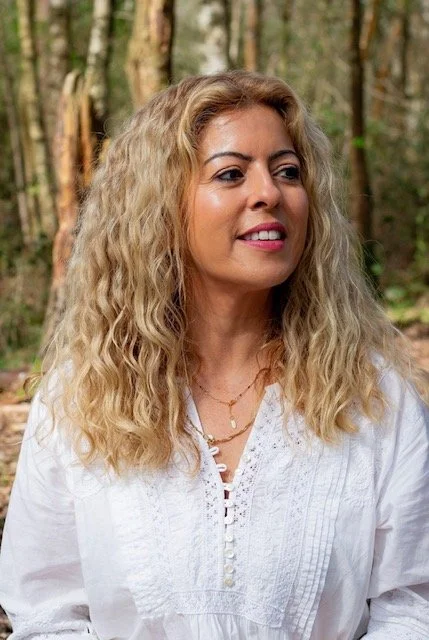 Woman with curly blonde hair outdoors in a forest, wearing a white blouse and layered necklaces.