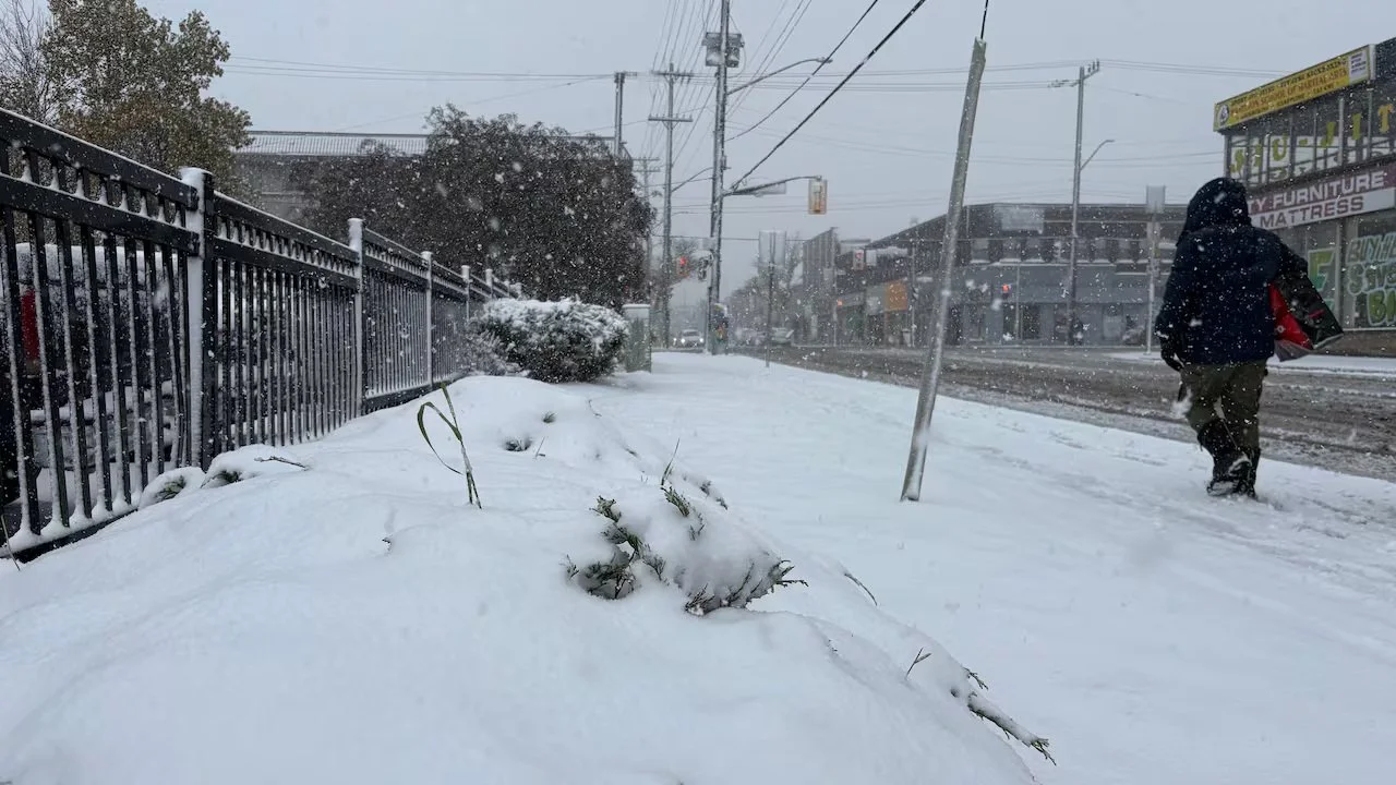 Snow-covered sidewalk with a person walking away, buildings and power poles in the background, snow falling.