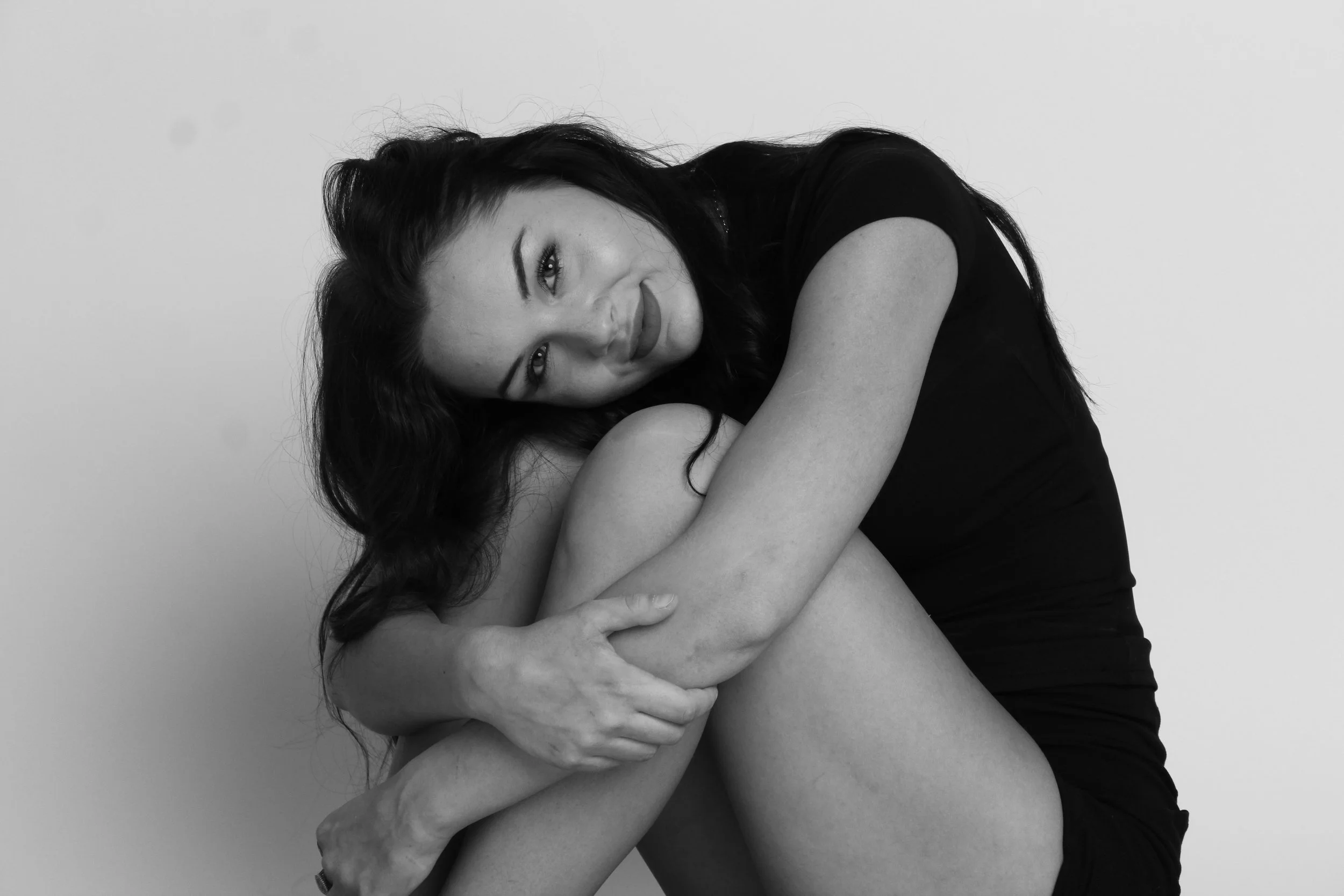 A woman with long dark hair sitting with her arms wrapped around her knee, smiling softly, wearing a black top against a plain background.