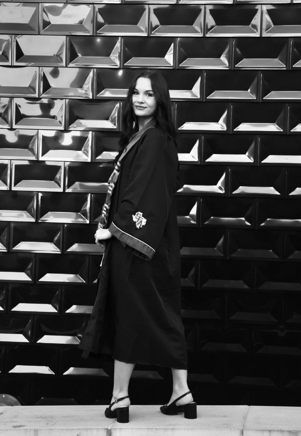 A woman with long dark hair wearing a black graduation gown and heels stands on a sidewalk in front of a decorative wall with geometric metallic tiles.