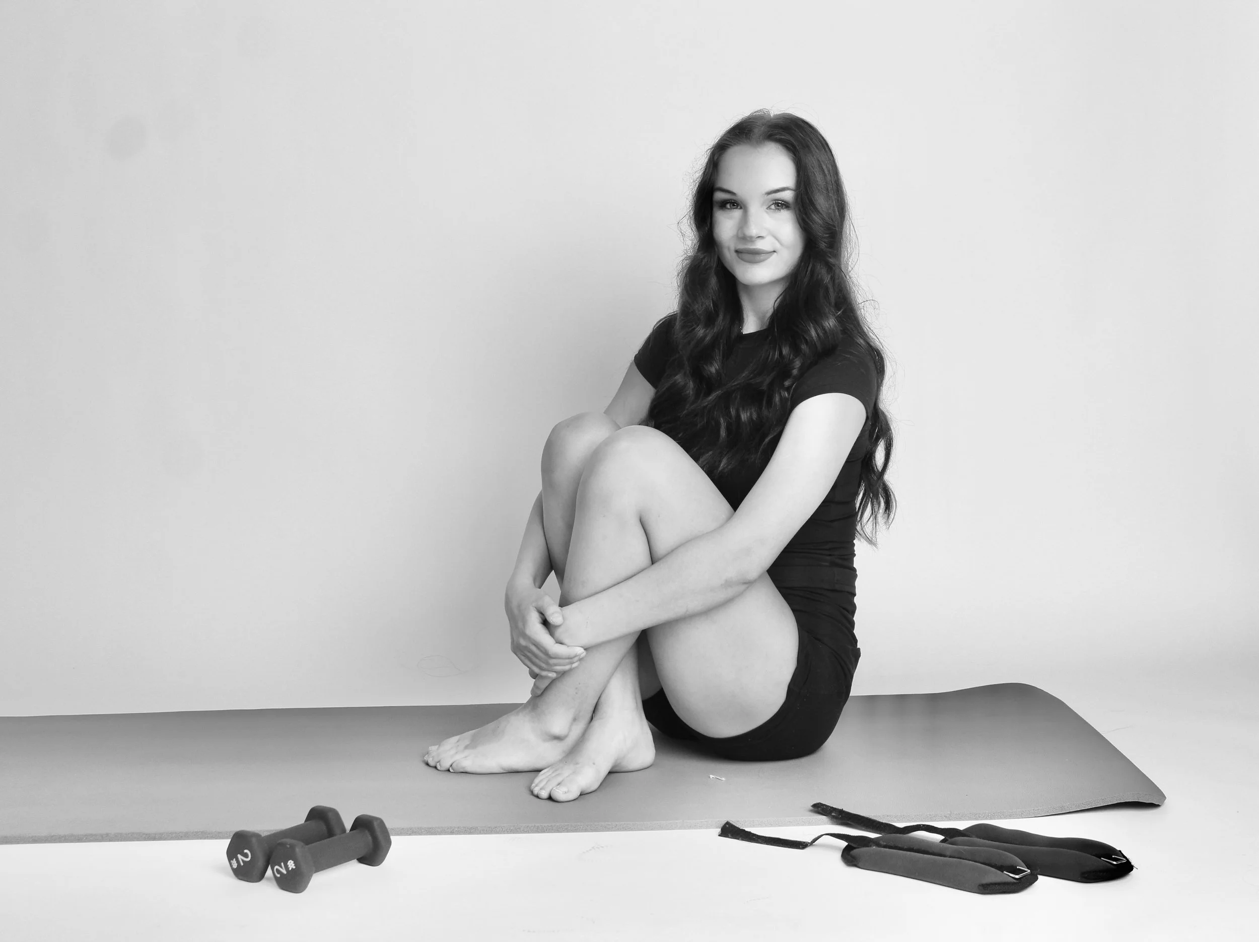 A young woman seated on a yoga mat with her knees bent and arms wrapped around her legs, smiling at the camera. She has long, dark, wavy hair and is wearing a black t-shirt and shorts. There are two small dumbbells and resistance bands on the floor near her, and the background is plain and light-colored.