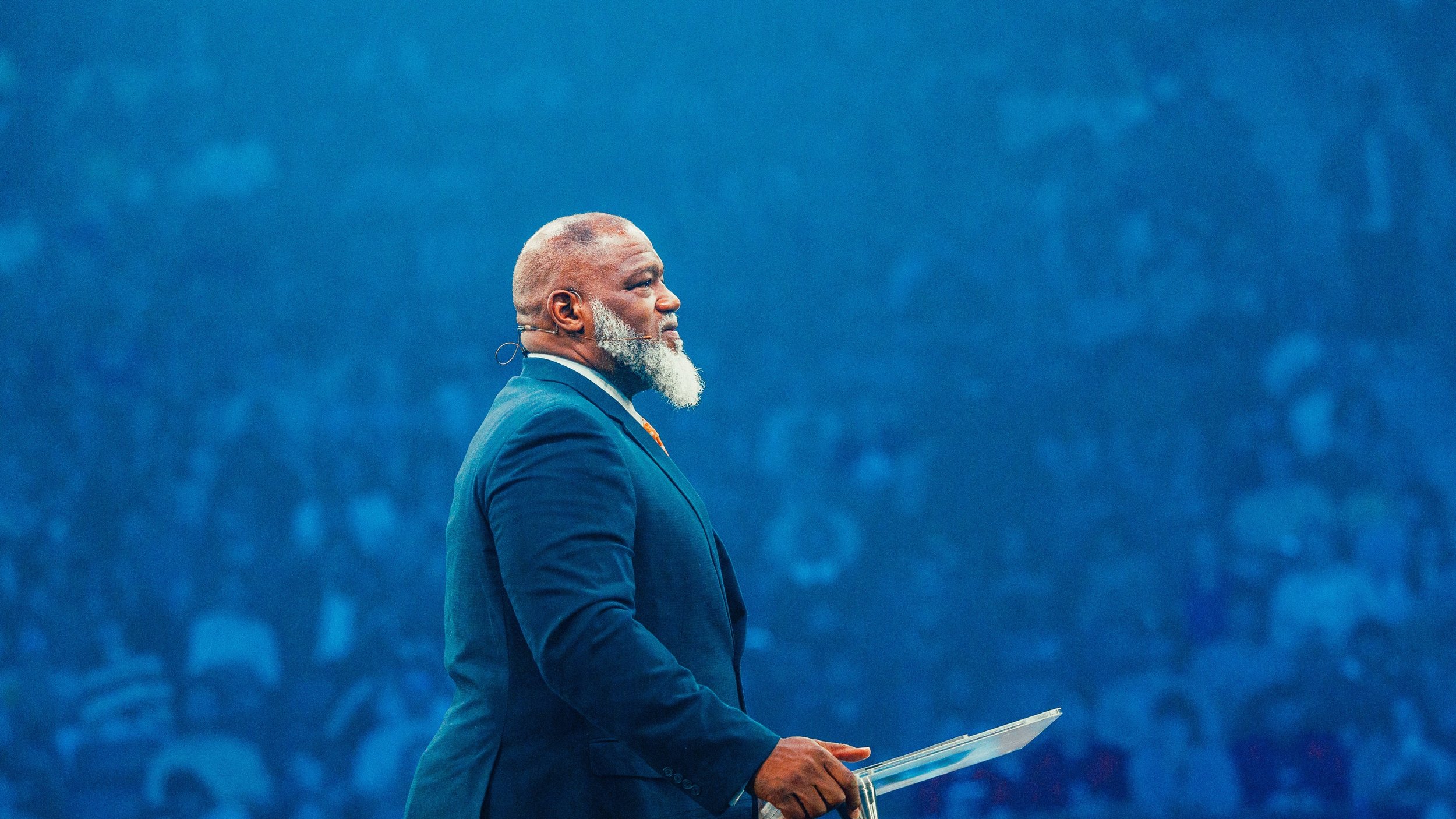 A man with a gray beard and short gray hair, wearing a dark blue suit, is standing on a stage holding a clear acrylic podium. The background is blurred with a large audience and blue lighting.