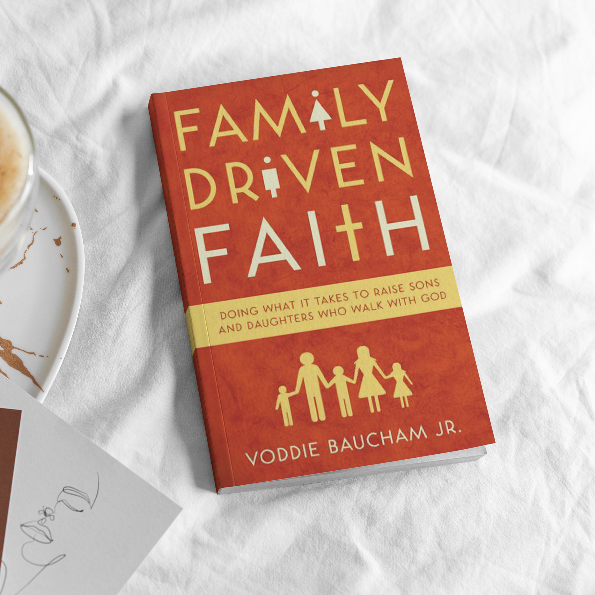 Book titled "Family Driven Faith" by Voddie Baucham Jr., lying on white fabric with a cup of coffee on a saucer partially visible on the left.