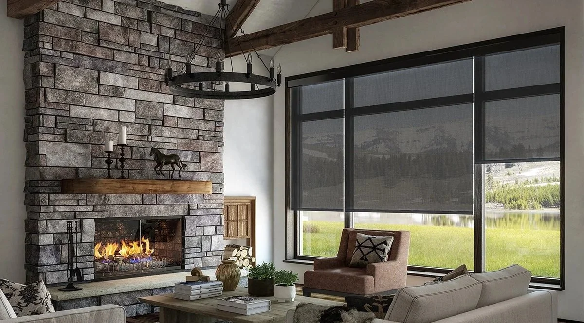 Living room with stone fireplace, wooden mantel, and large windows with shades overlooking a lake and mountains.