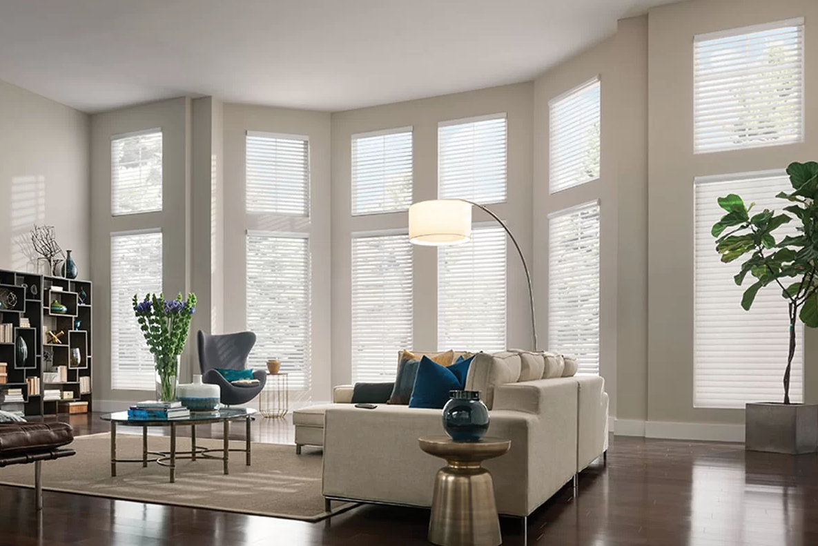 Living room with large bay windows, a white sofa with blue pillows, a tall floor lamp, a black bookshelf, a glass coffee table with a vase of purple flowers, and hardwood floors.