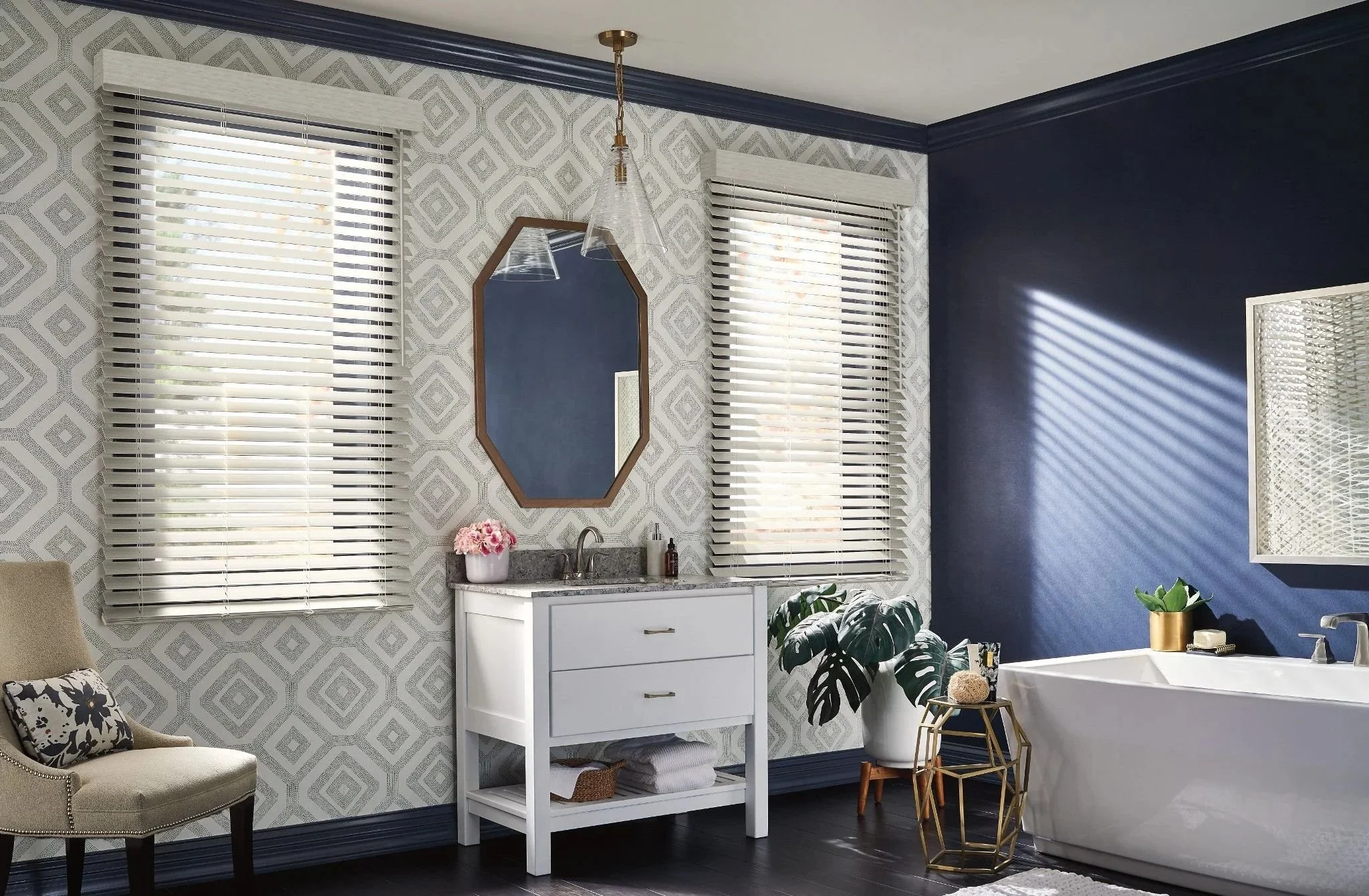 Bright bathroom with navy and patterned wallpaper, two windows with white blinds, a white vanity with a granite top, a mirror, a bathtub, and decorative plants and accessories.