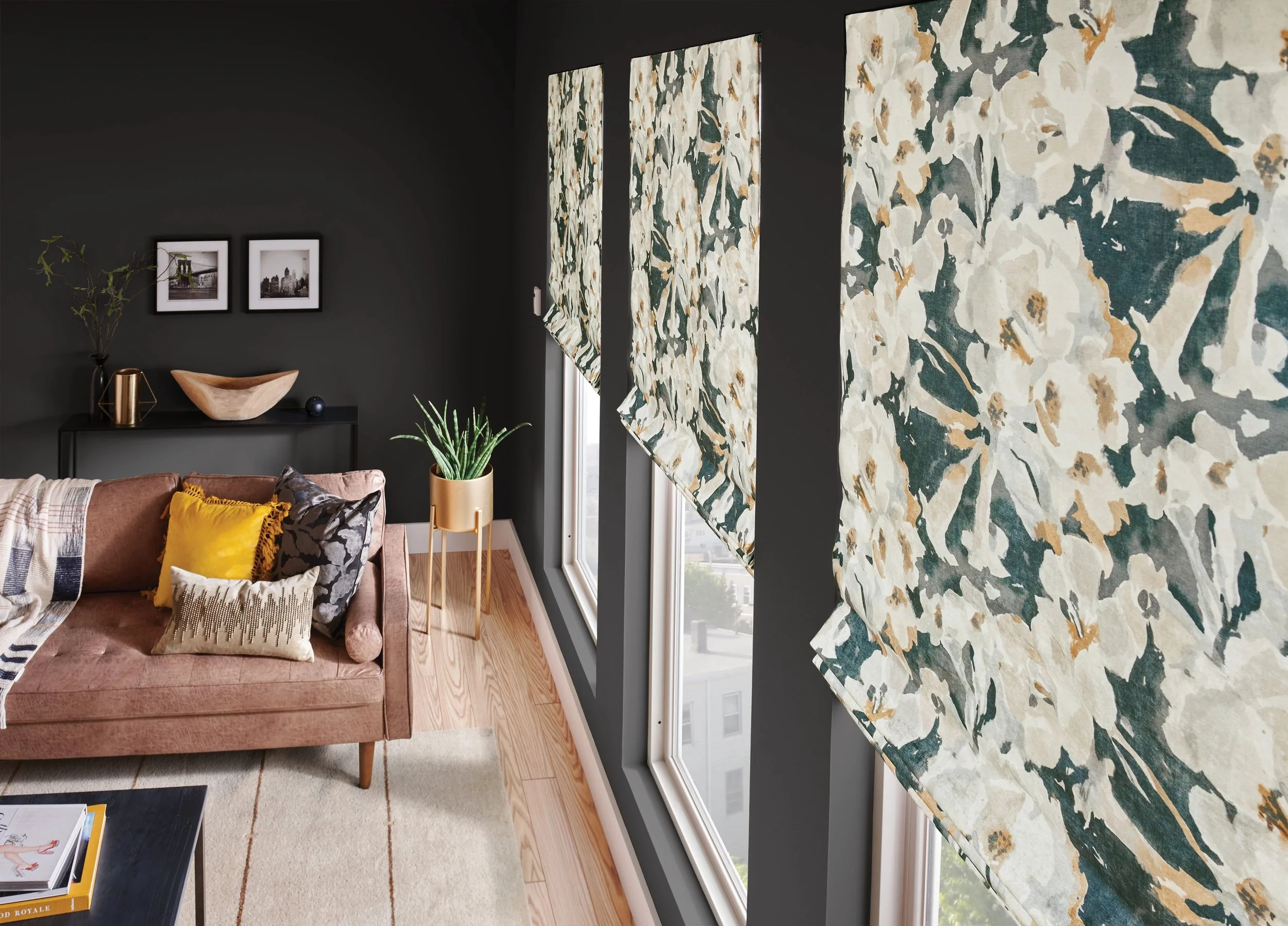 Living room with large windows covered by floral-patterned blinds, dark painted walls, a pink sofa with colorful pillows, a black sideboard with decorative items, a small potted plant on a stand, and framed black-and-white photographs on the wall.