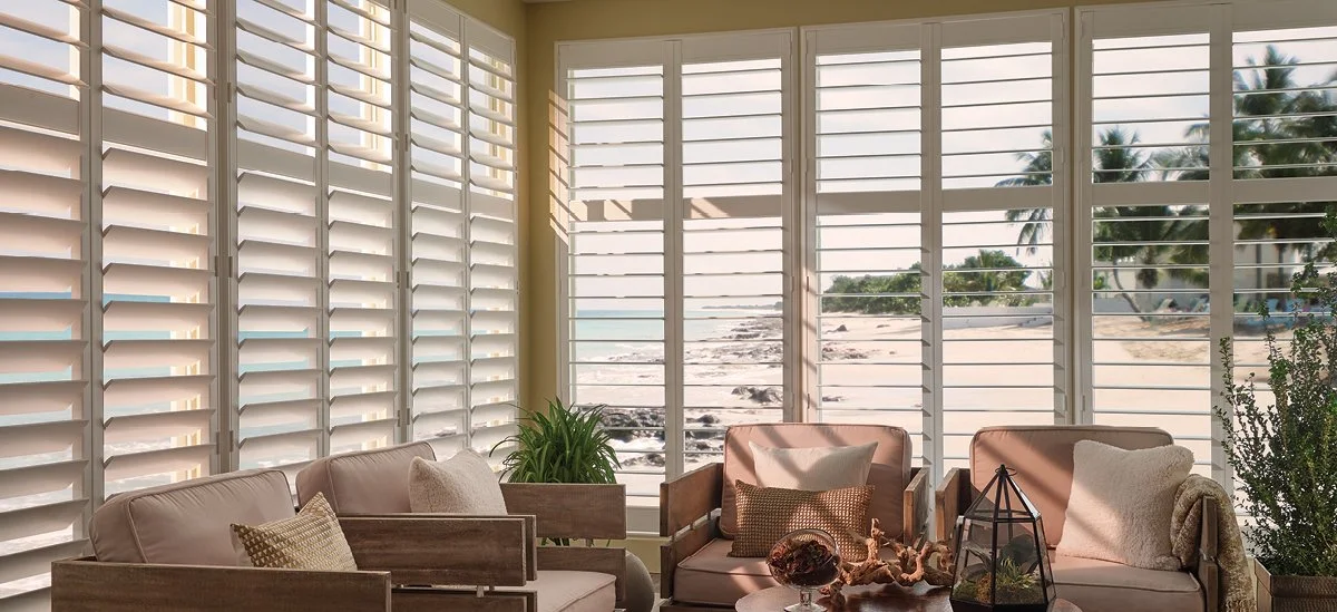 Living room with large windows and white plantation shutters, overlooking a beach with palm trees.