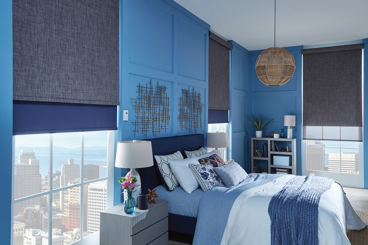 Modern bedroom with blue walls, large windows with city view, and dark gray and blue window treatments. The bed has white and patterned pillows, a navy upholstered headboard, and is covered with a light blue quilt and a darker blue throw. There are two nightstands with table lamps and decorative items, and a small bookshelf with plants and decor.