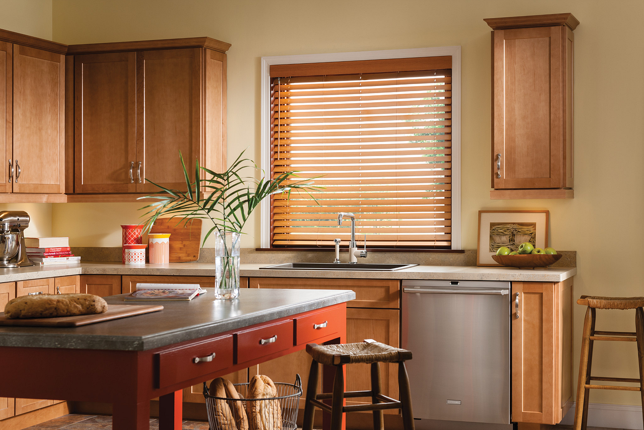 A kitchen with wooden cabinets, a window with blinds above the sink, a red kitchen island with a cutting board and bread, a potted plant, and a basket of bread on the floor.