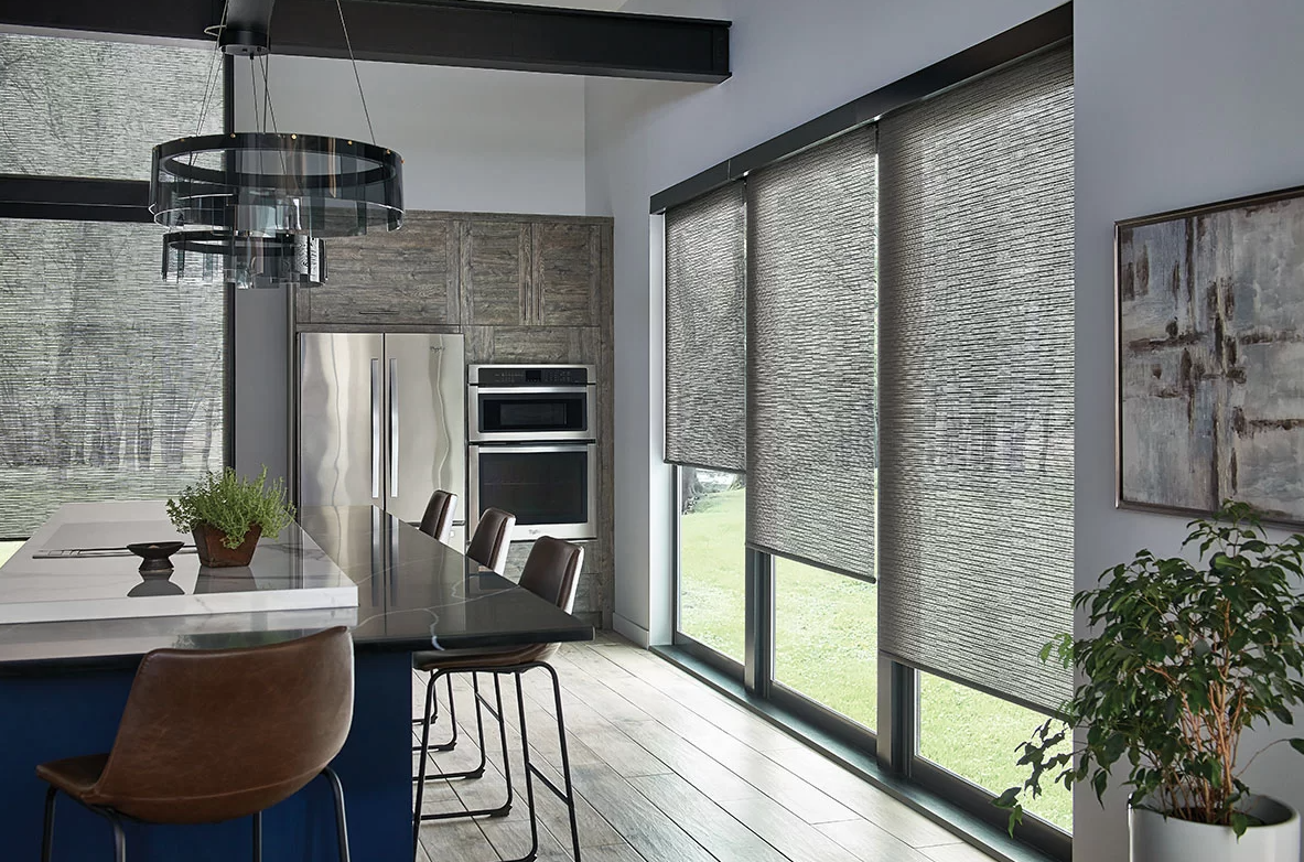 Modern kitchen with large windows covered by textured roller blinds, a black kitchen island with a white countertop, and stainless steel appliances, including a double oven and a refrigerator, with paper white walls and a wooden floor.
