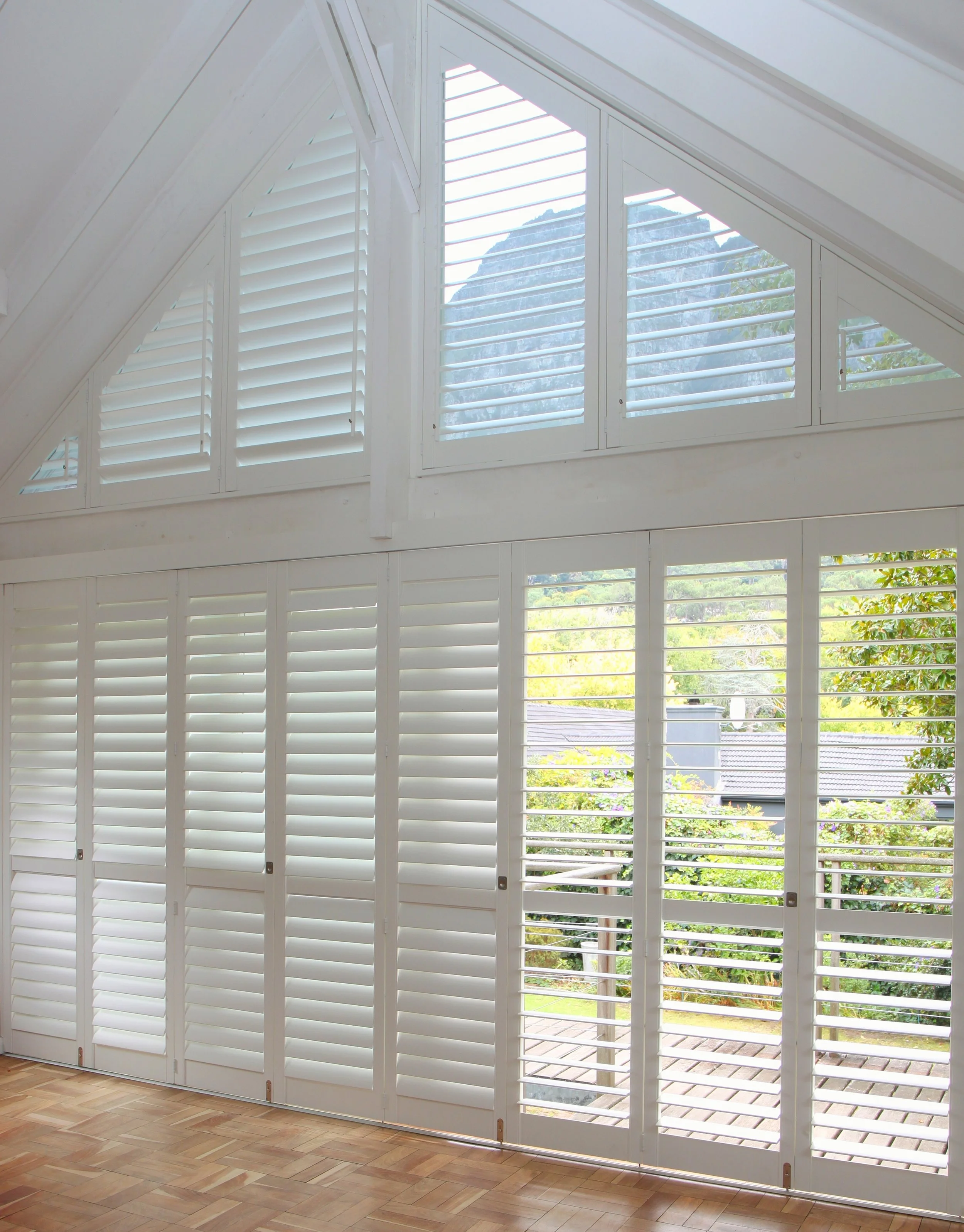 Interior view of a room with large white plantation shutters covering windows, showing a view of a mountain and garden outside.