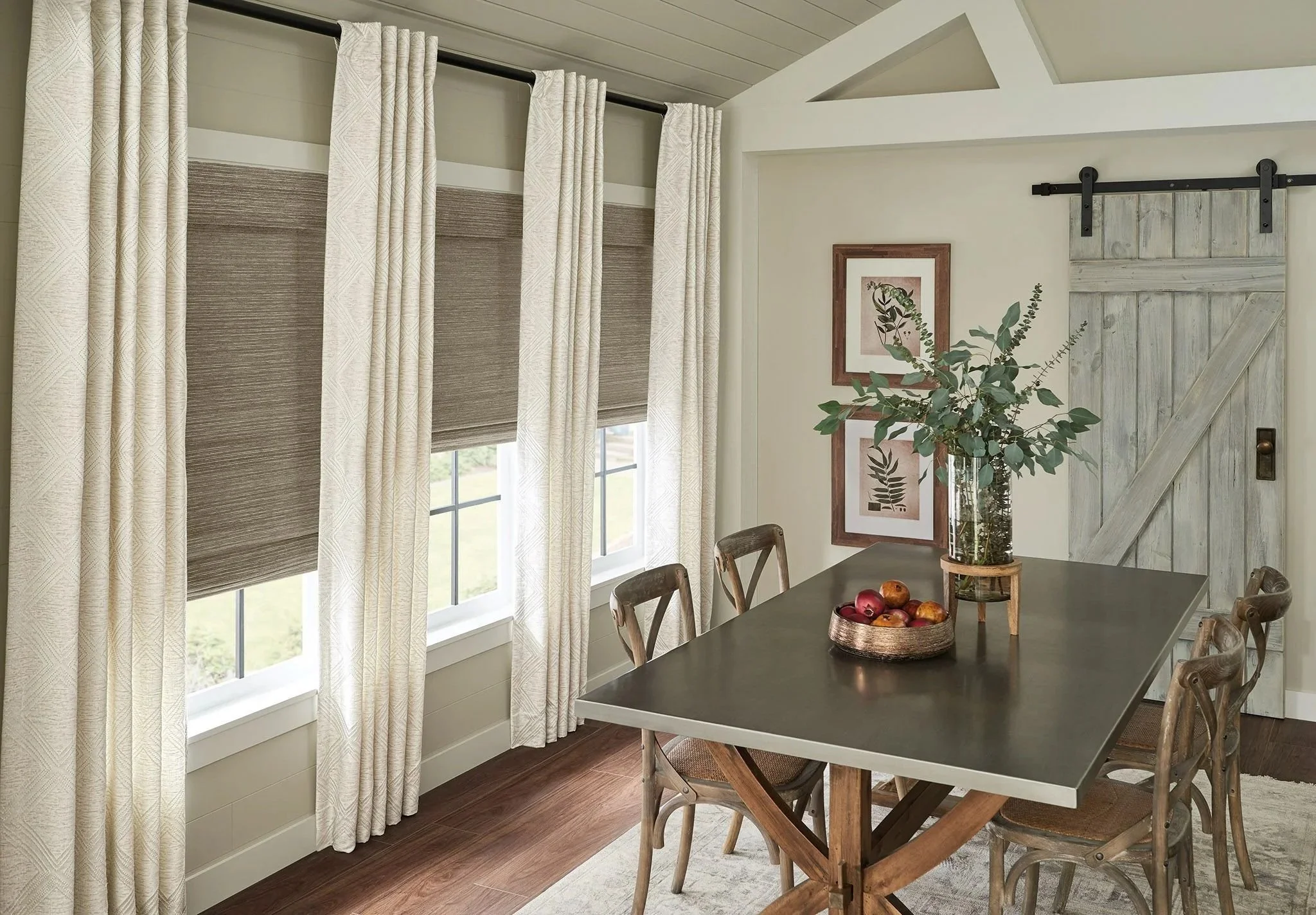 A dining room with large windows covered by beige curtains and brown shades, a wooden table with six chairs, a glass vase with green foliage, and framed botanical prints on the wall, along with a sliding barn door.