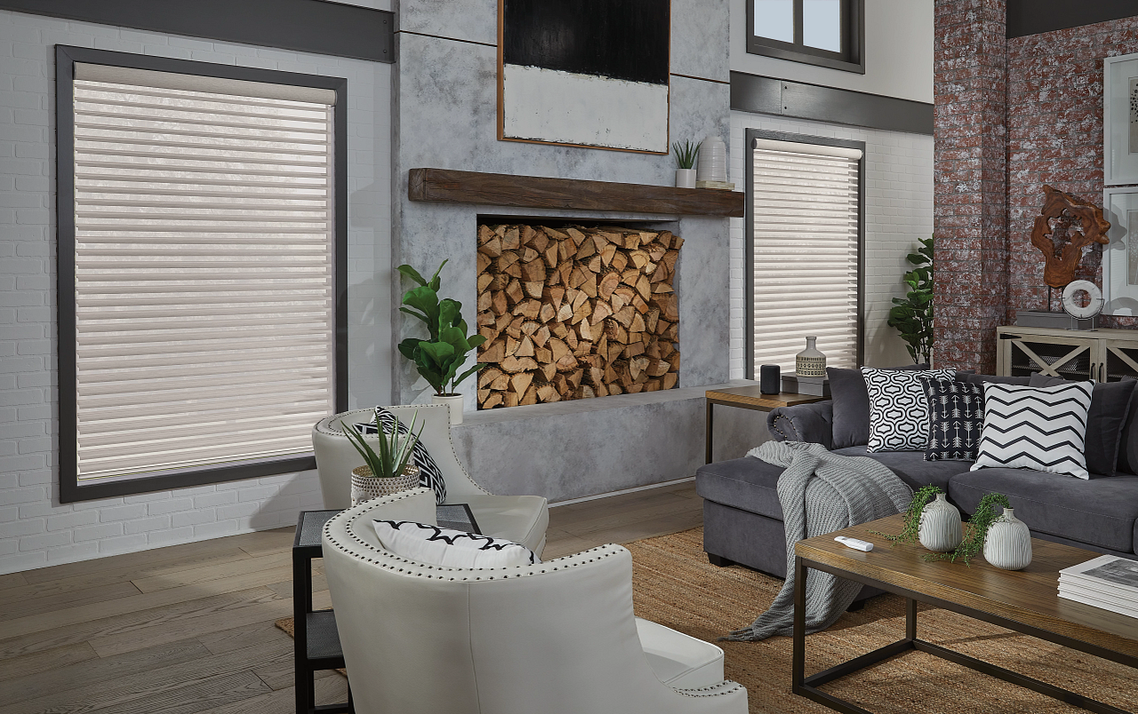 Living room with white brick and textured walls, wood fireplace with stacked firewood, two large windows with white blinds, gray sofa with black and white patterned pillows, white armchairs, wooden coffee table with white vases, potted plants, and decorative items.