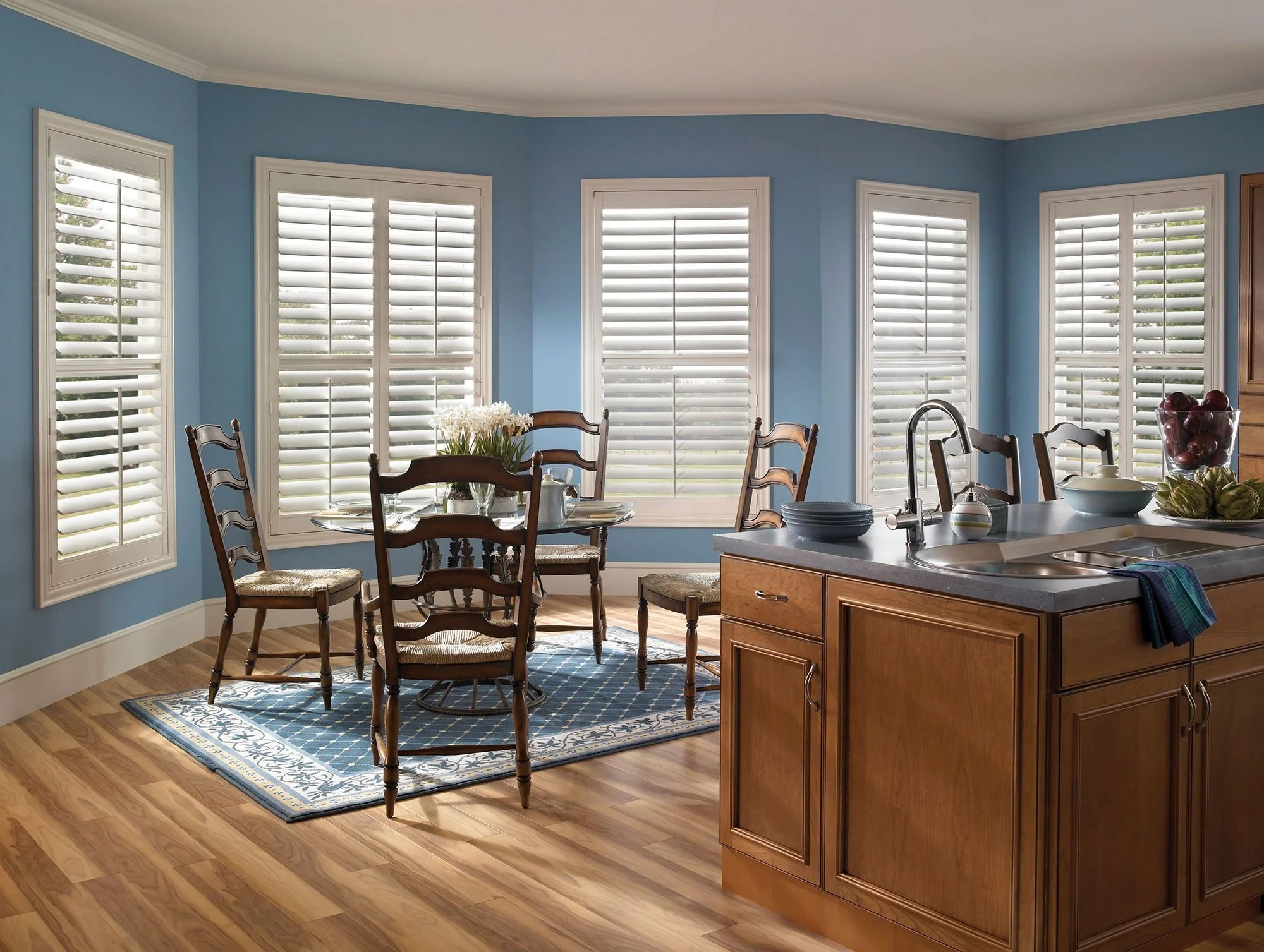 A bright kitchen and dining area with blue walls, hardwood floors, a round wooden dining table with six chairs, and multiple large windows with white plantation shutters. The kitchen features a wooden island with a sink, bowls, and a towel.
