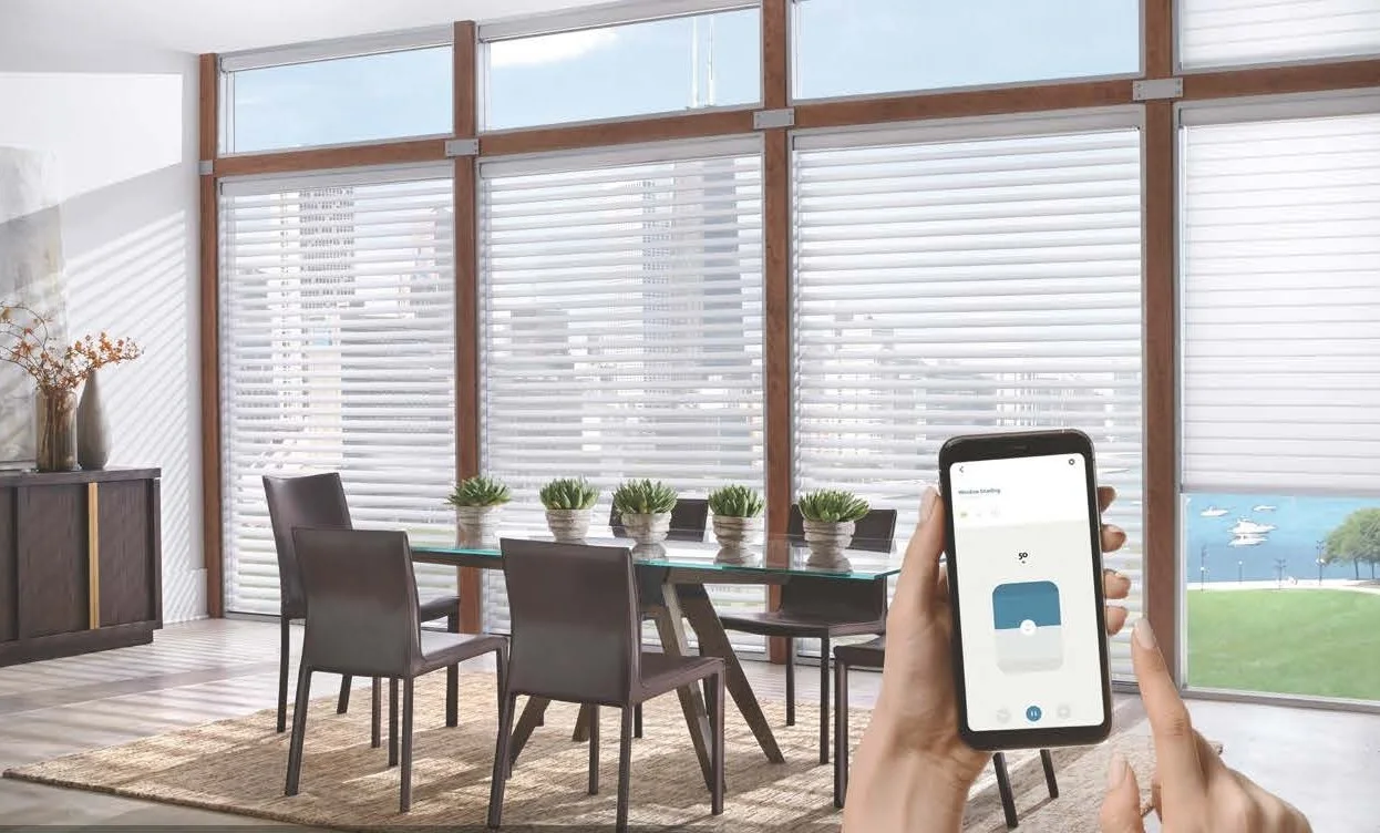 Interior of a bright dining room with large windows and horizontal blinds. There is a glass dining table with six black chairs and a row of small potted plants on the table. A woman is holding a smartphone controlling window blinds.