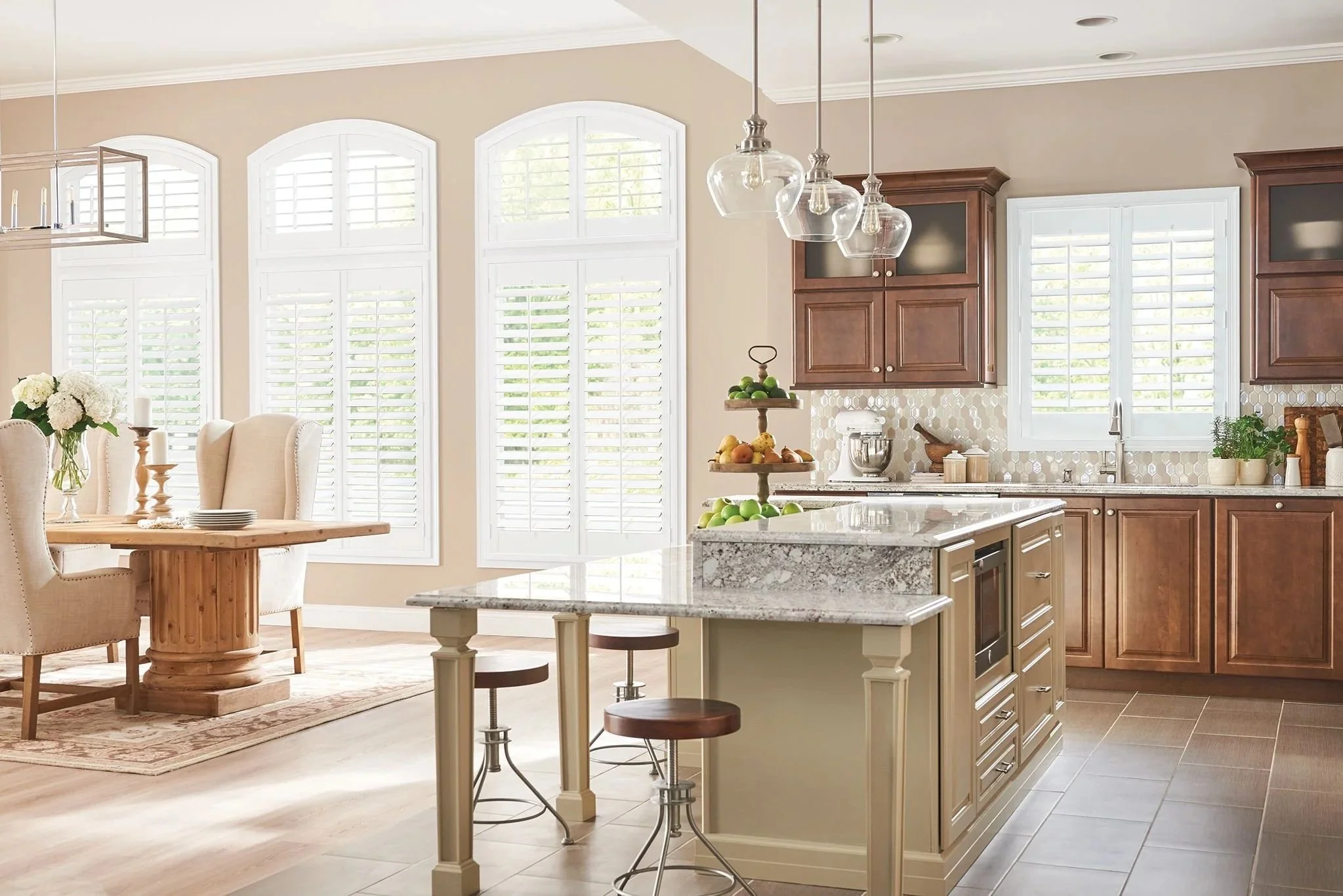 Bright kitchen with large windows, wooden cabinets, granite countertops, and a central island with fruit display, adjacent dining area with a wooden table and cream-colored chairs.