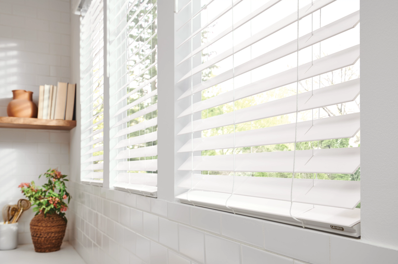 A bright kitchen window with white horizontal blinds, a white tiled wall, a wooden shelf with books and a brown vase, a woven basket with a plant, and a white container with kitchen utensils.