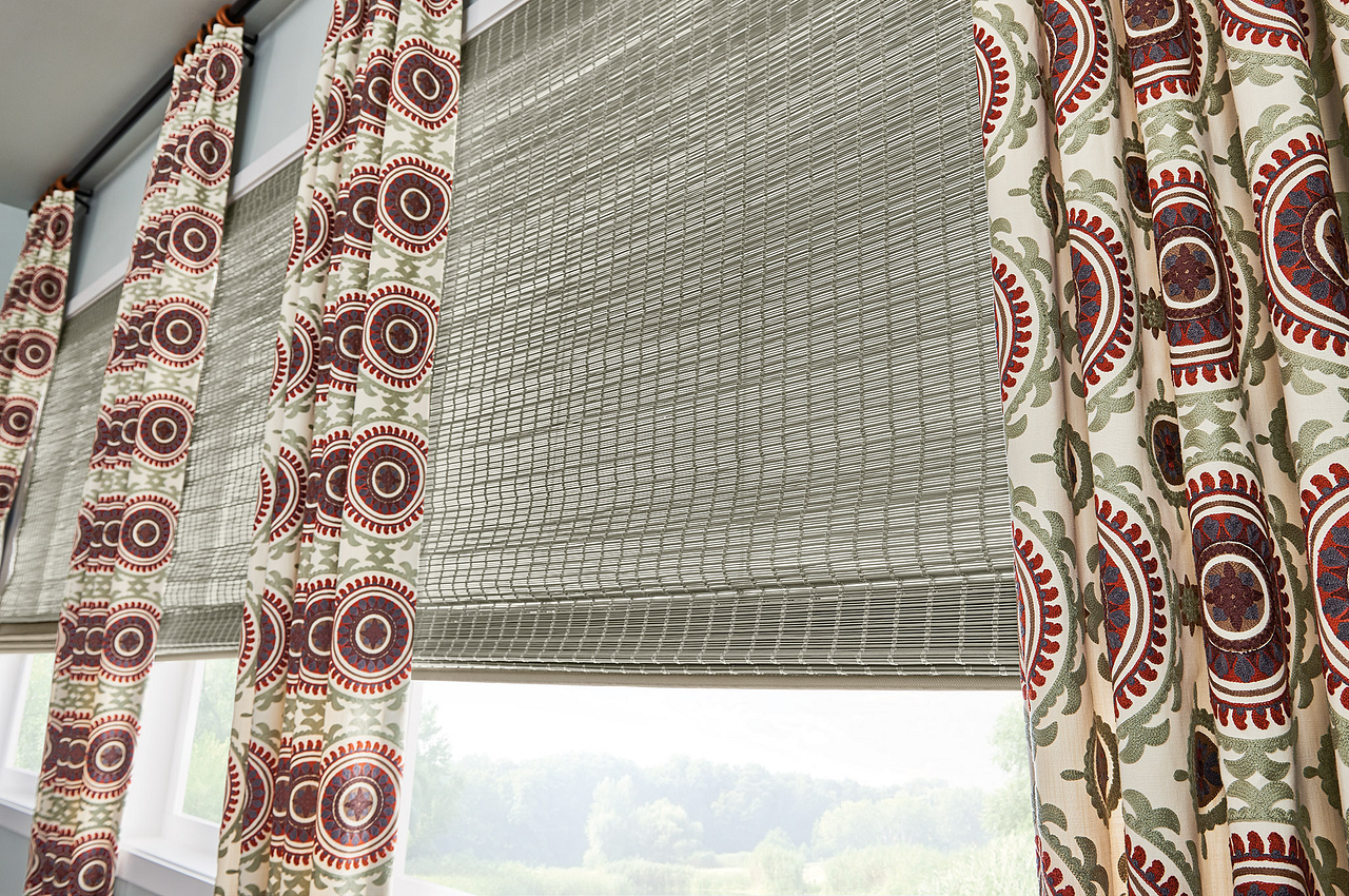 Window with patterned curtains and a woven shade, overlooking a green landscape.