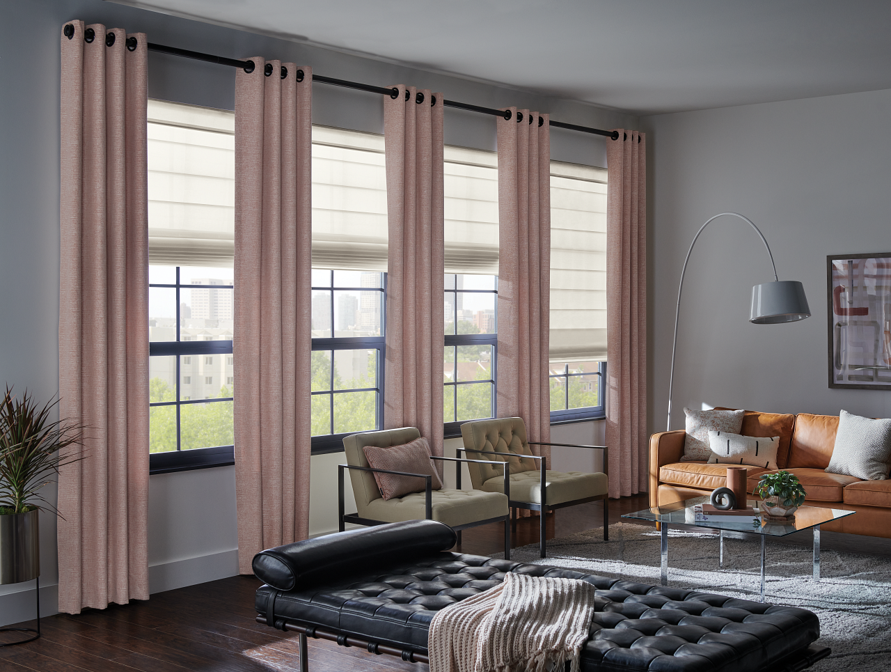 Modern living room with large windows, pink curtains, beige and gray seating, a leather chaise lounge, a coffee table with decorative items, and a potted plant.