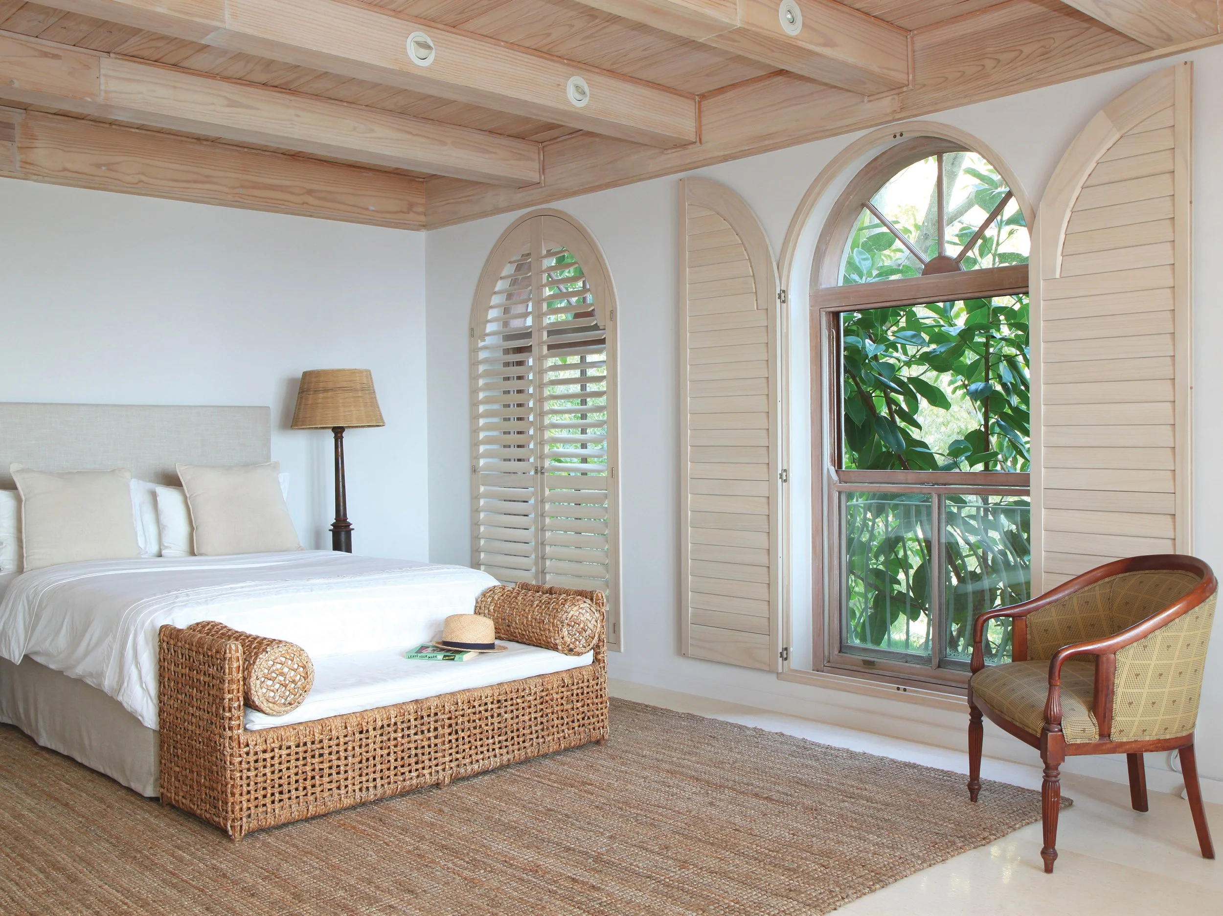 A bright bedroom with a bed, wicker bench, lamps, and large windows with wooden shutters overlooking greenery.