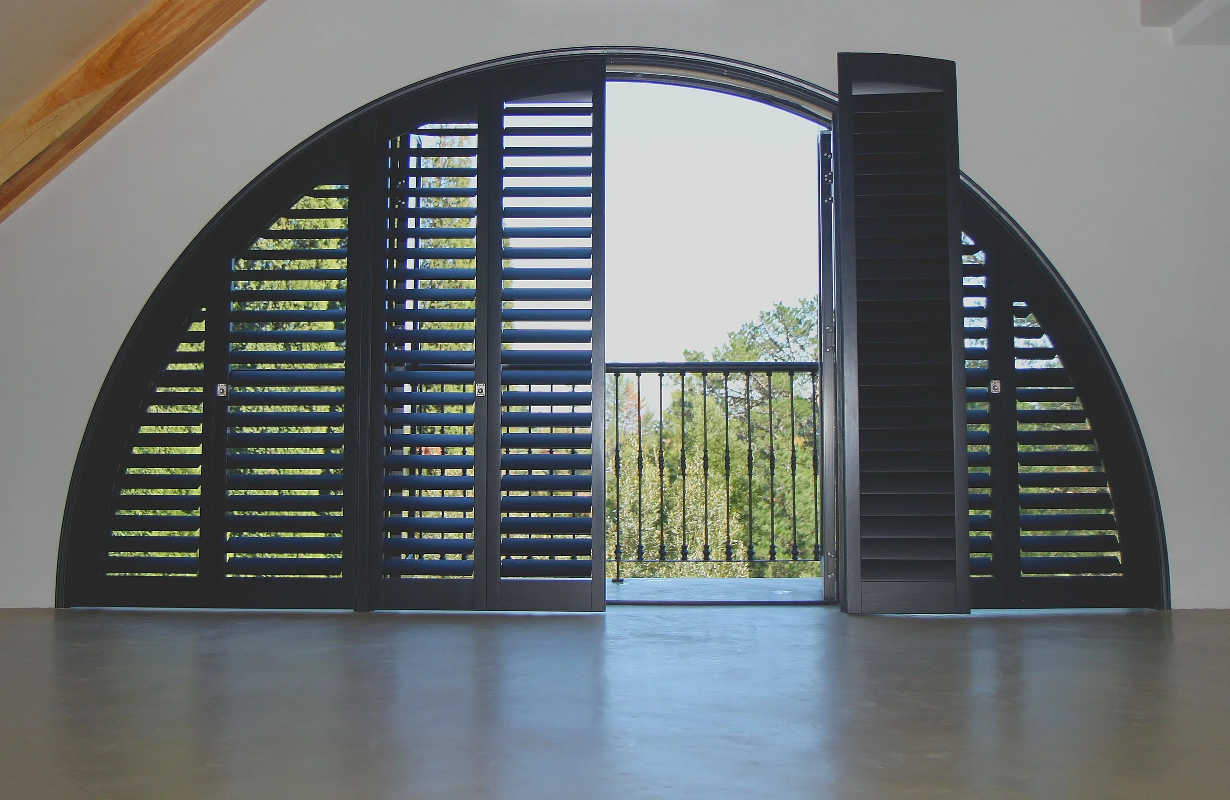 Open black louvered shutters revealing a small balcony with a black railing and a view of green trees outside.