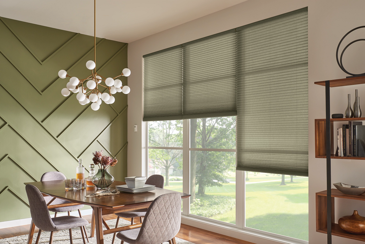 Dining room with green accent wall, wooden table set with dishes and vases, large window with beige blinds, and a shelving unit with books and decorative items.