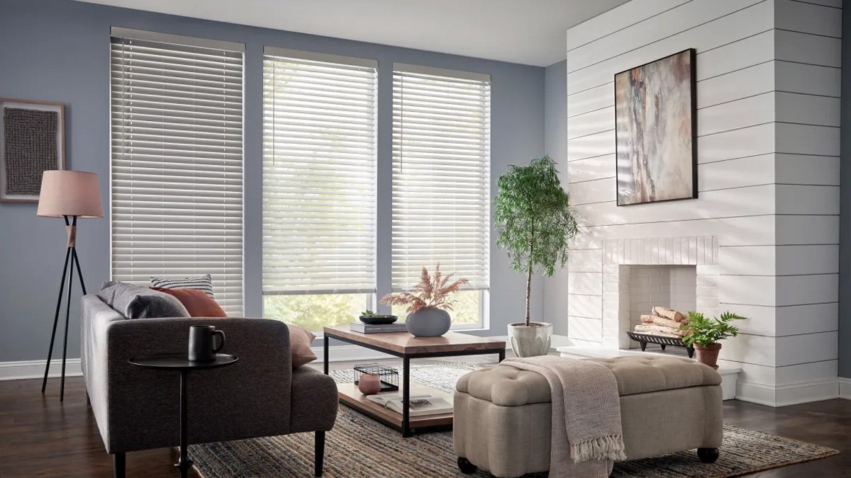 Living room with large windows, white blinds, gray sofa with pillows, small round side table with a mug, wooden coffee table with a plant and decorative objects, fireplace, artwork on the white paneled wall, indoor plants, and a patterned rug.
