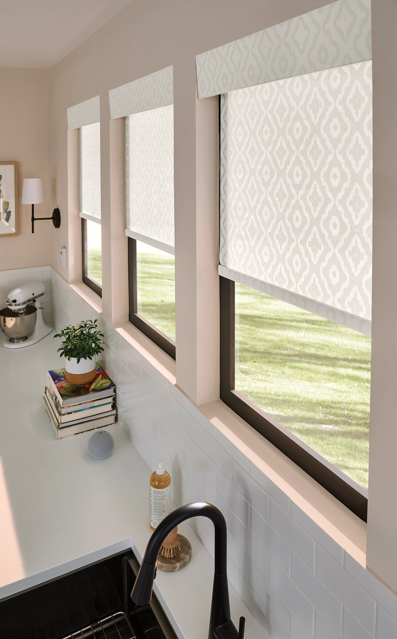 Three windows with patterned roller shades in a kitchen, with a plant on a stack of magazines, a soap dispenser, a sink faucet, and a stand mixer on the countertop.