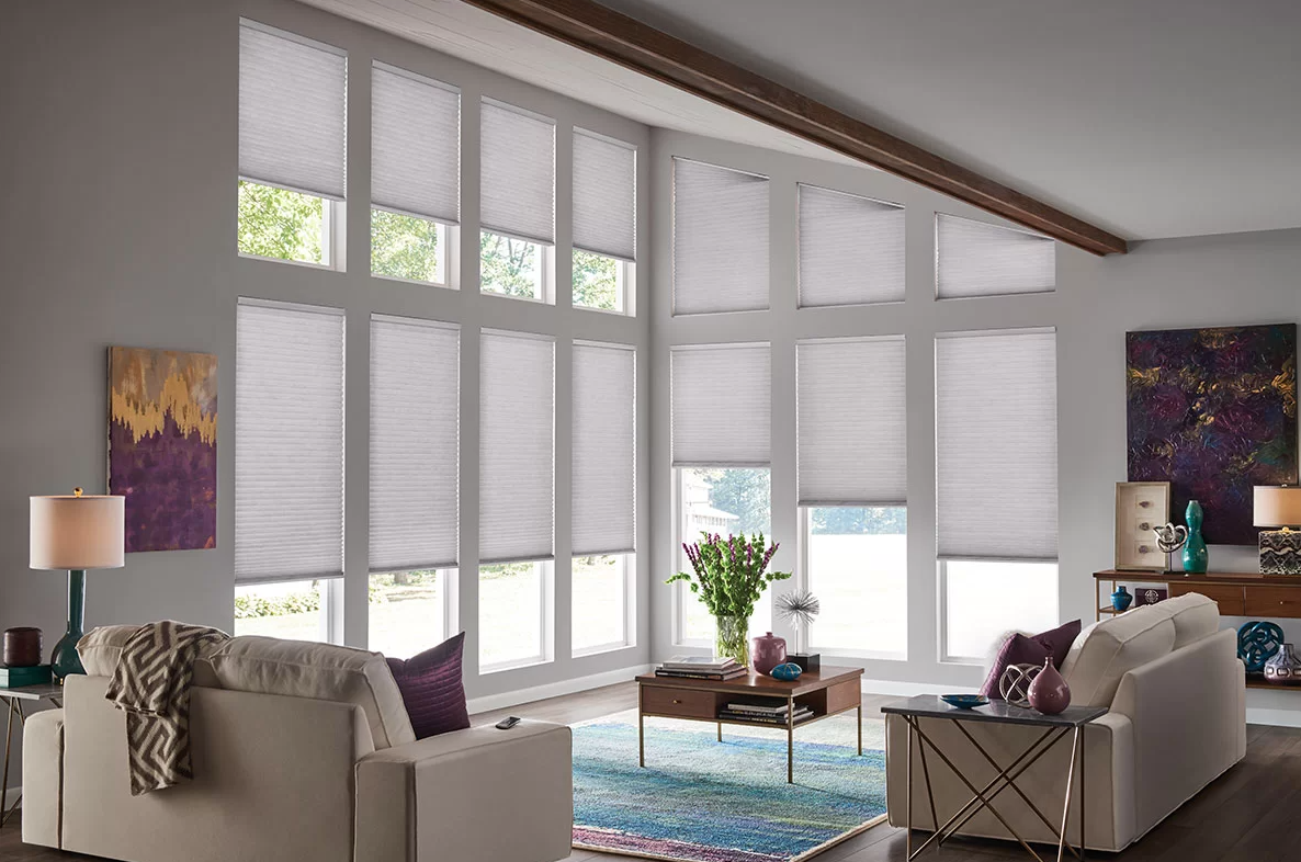 Bright living room with large windows covered by white blinds, white sofas with purple pillows, a colorful area rug, a wooden coffee table with a vase of flowers and decorative objects, and abstract artwork on the walls.