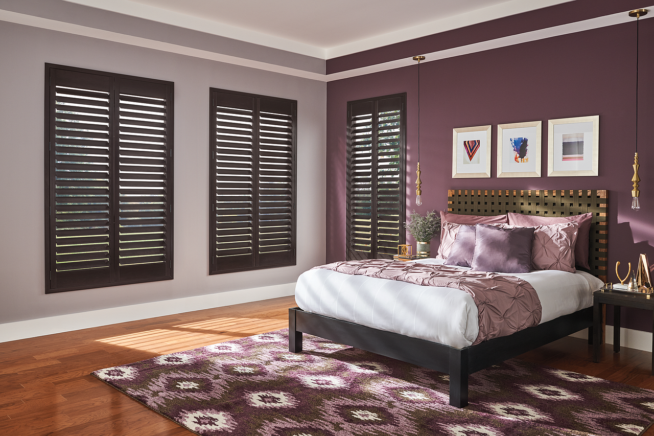 A modern bedroom with a purple accent wall, black window shutters, a wooden bed with purple and white bedding, a patterned rug, and framed artwork above the headboard. Two hanging pendant lights are on either side of the bed.