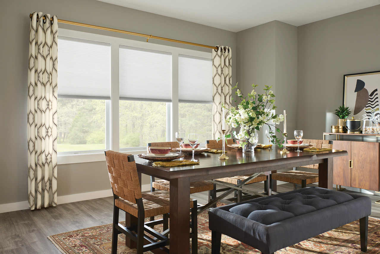 A dining room with a wooden table set for a meal, including wine glasses, plates, and gold flatware. There is a large floral centerpiece, a black upholstered bench, and four woven chairs. The room has three large windows with white roller blinds and patterned curtains, with a sideboard featuring gold accents and decorative items. The floor is wood with a patterned rug.