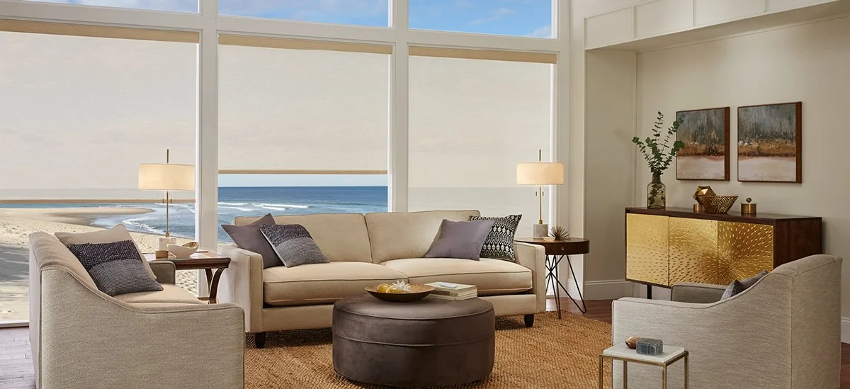 Living room with large windows overlooking the beach, beige sofas, armchairs, lamps, side tables, wall art, and a sideboard, with a view of the ocean outside.