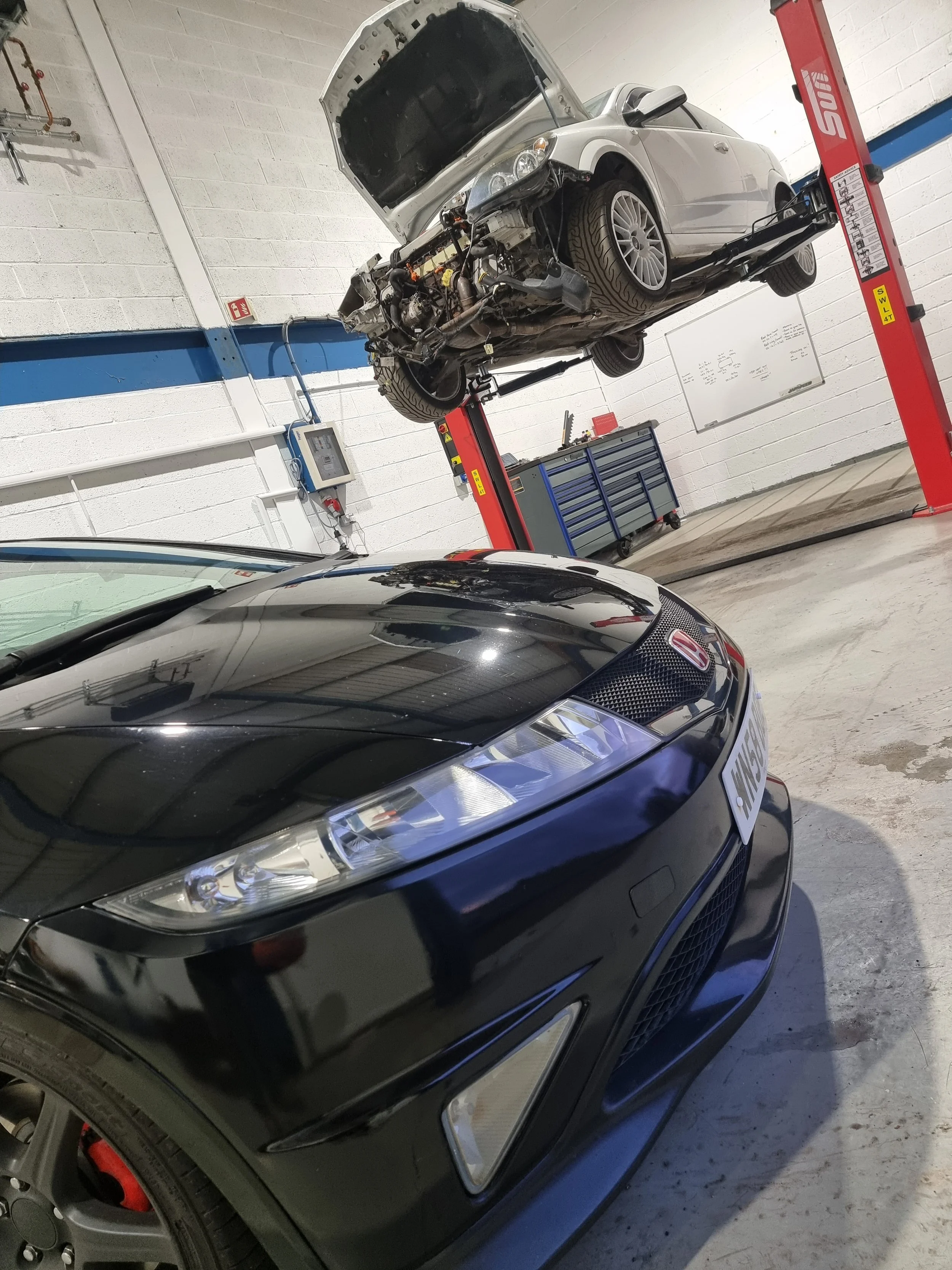 Inside the workshop with two cars: a black Honda Civic Type R in the foreground and a white Vauxhall Astra VXR on a lift in the background, which is undergoing repairs to the exhaust manifold studs and a new turbo.