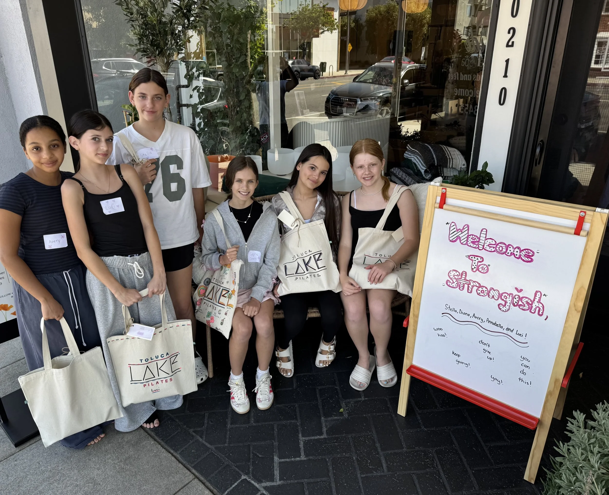 Group of young girls standing outside a storefront with large glass windows, holding tote bags, and smiling at the camera. There is a whiteboard sign beside them with colorful writing.